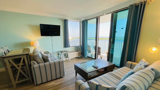 Ein Wohnzimmer mit Sofa, Couchtisch und Fernseher. Ein Balkon bietet Blick auf das Meer.