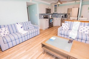 A living room with two striped sofas, a coffee table, and a kitchen area with white cabinets and a microwave.