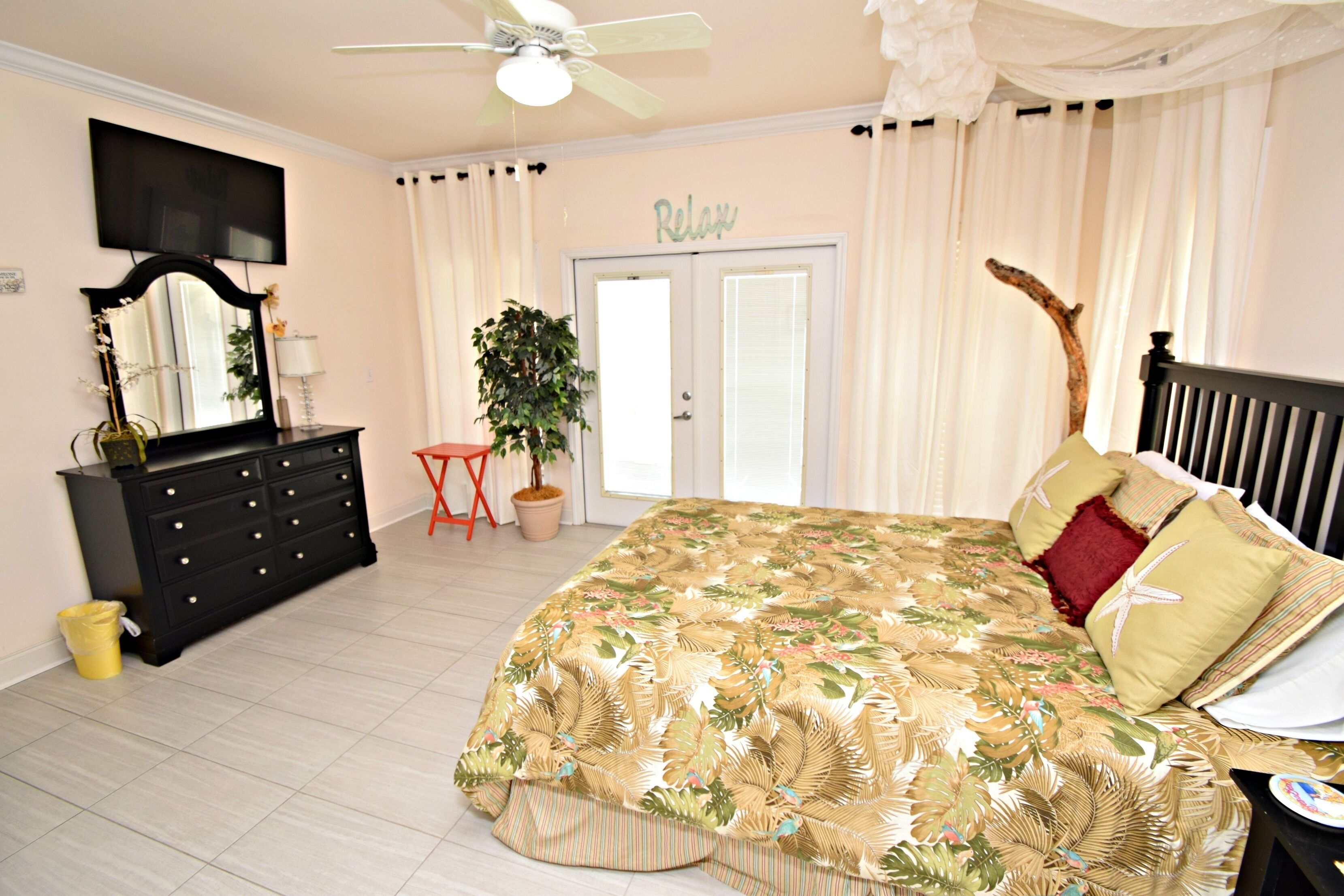 A bedroom with a bed, a dresser, a mirror, a television, a ceiling fan, and a door with a 'Relax' sign.