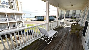 Une terrasse en bois avec une balustrade blanche, des chaises longues et une vue sur la plage et les bâtiments.