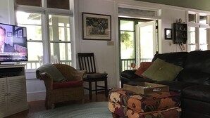 Een woonkamer met een televisie, een rieten stoel, een zwarte leren bank en een gestreept voetenbankje.