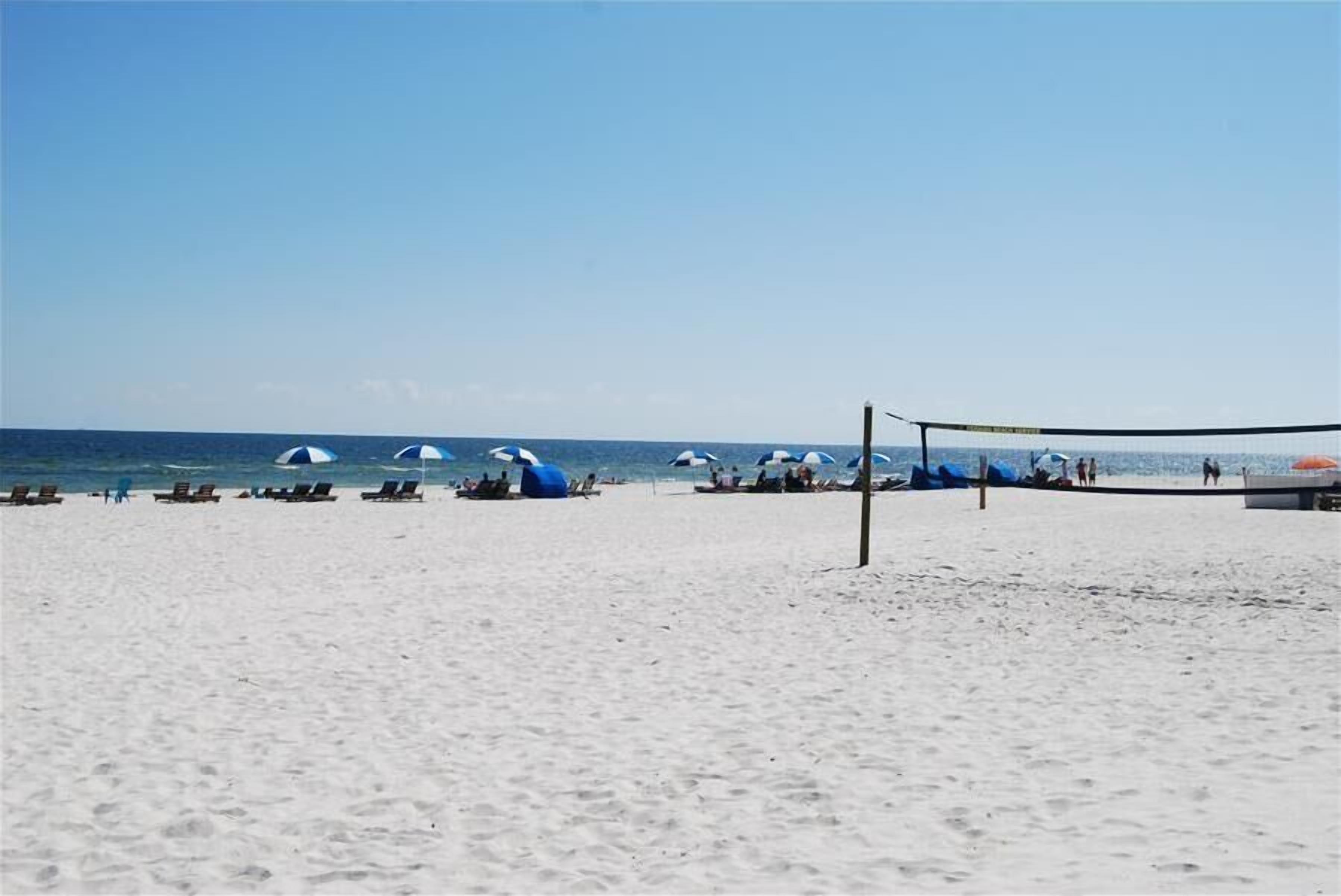 A beach with white sand, a volleyball net, and several umbrellas.