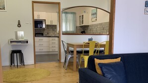 A living room with a sofa, a dining table with chairs, a kitchen area with cabinets and a microwave, and a small stool. - Dunes Beach House Goolwa Beach (Goolwa)