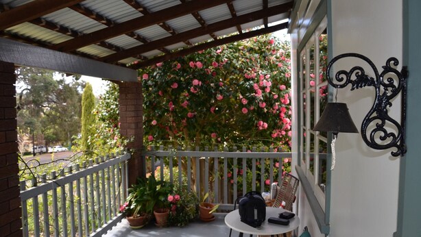 A covered balcony with a white railing, potted plants, a table with a chair, and a decorative wall-mounted light fixture.