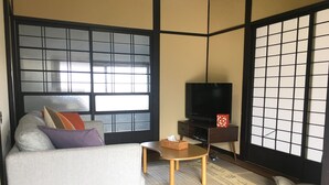 Ein traditioneller japanischer Raum mit Tatami-Böden, einem niedrigen Tisch, einer Couch mit Kissen, einem Fernsehgerät auf einem Schrank und verschiebbaren Shoji-Schiebetüren.
