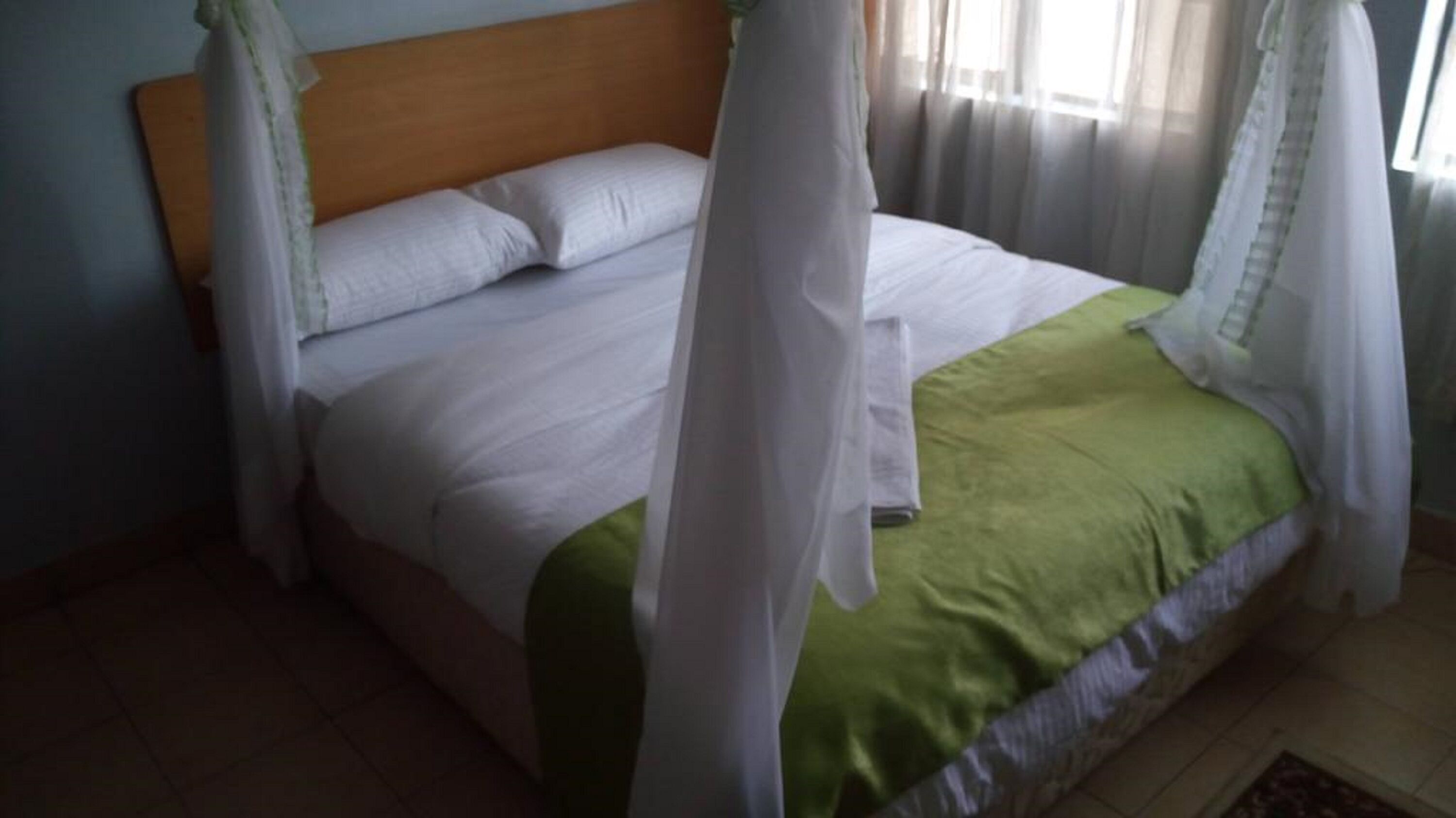 A bed with white bedding and a green blanket, a wooden headboard, and a window with sheer curtains.