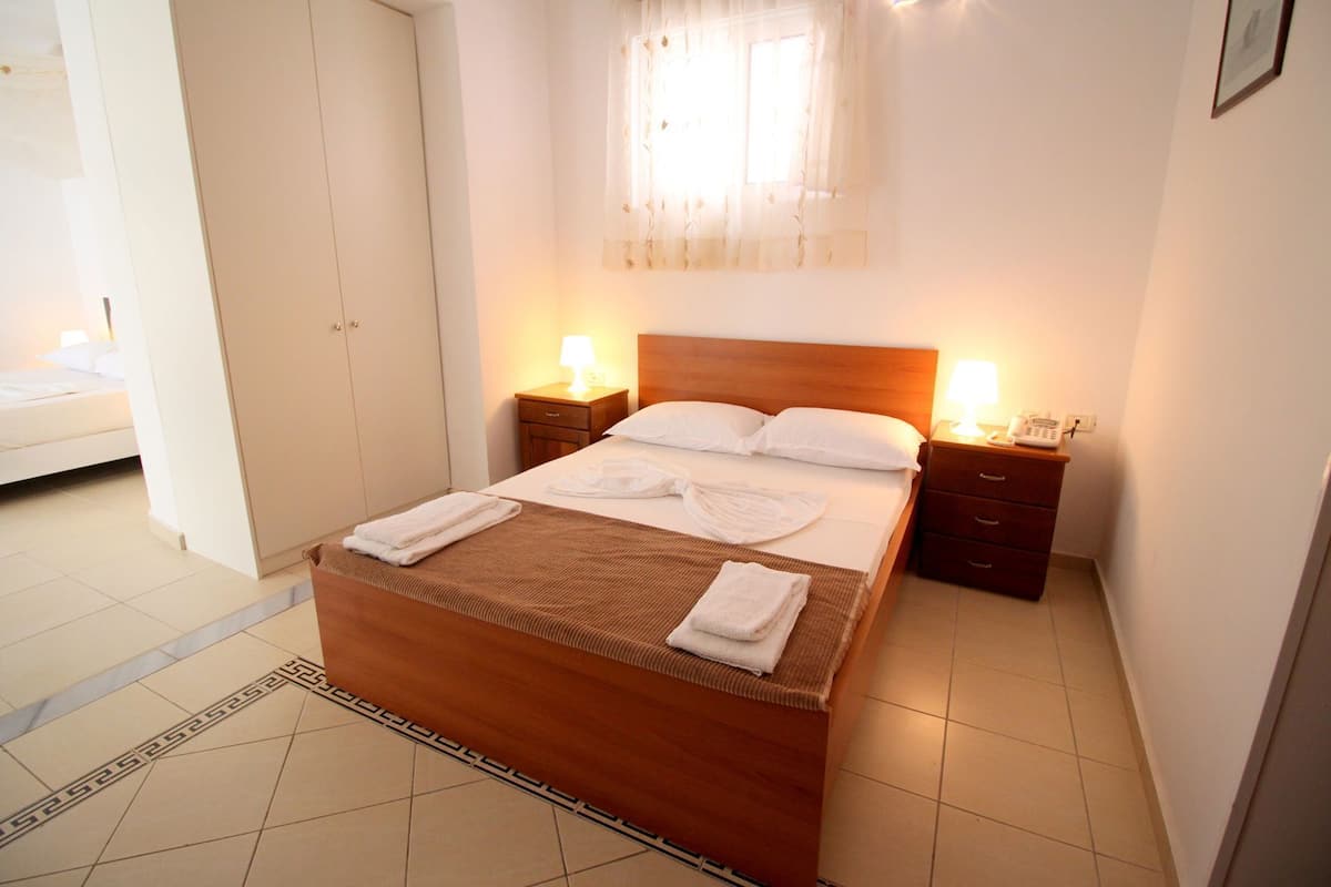 A hotel room with a wooden bed, two bedside tables with lamps, a window with sheer curtains, and a tiled floor.