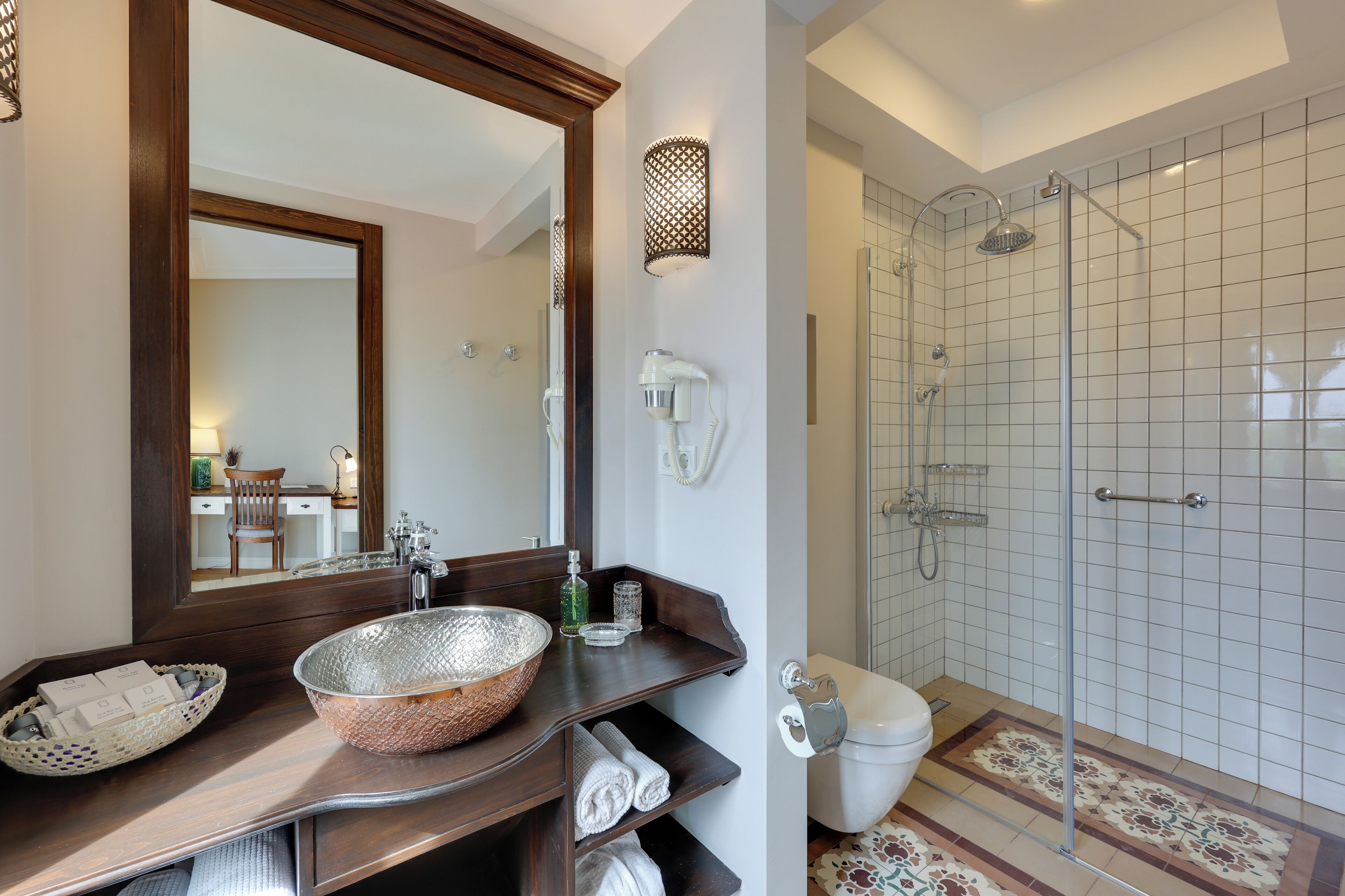 A bathroom with a large mirror, a sink with a woven bowl, a shower with a glass door, and a tiled floor.