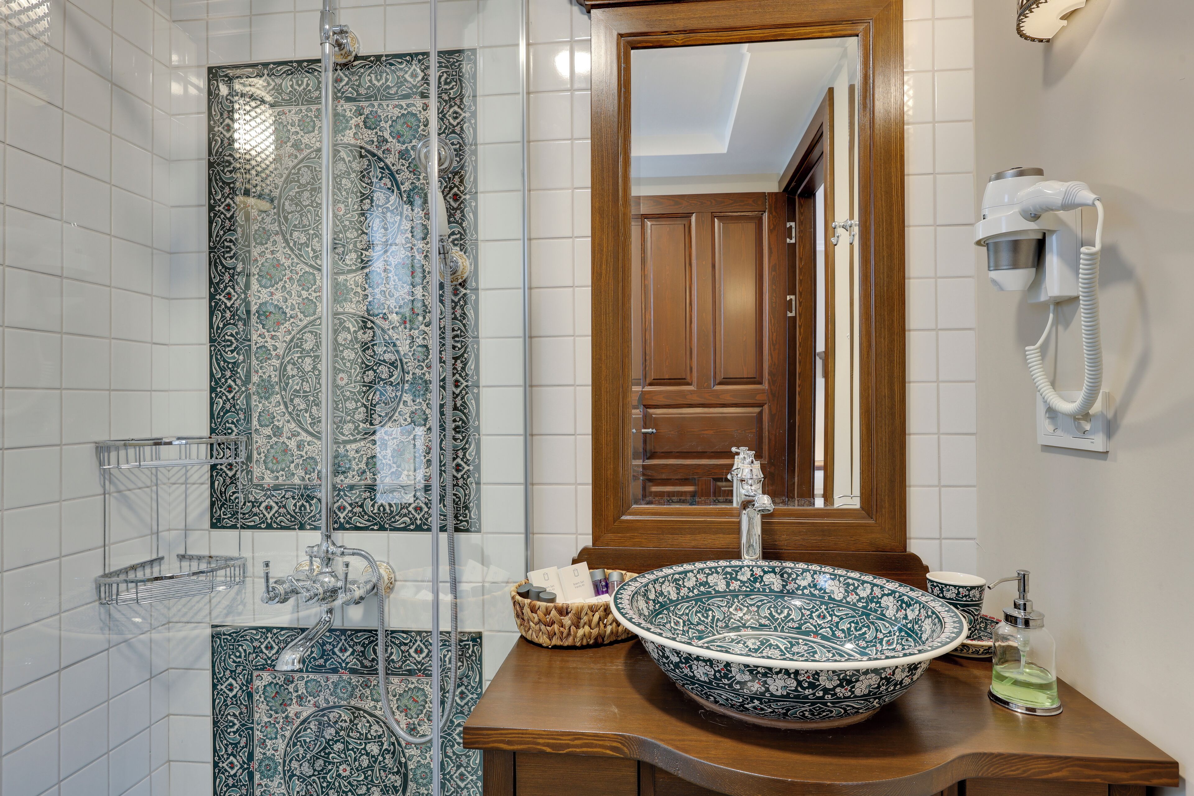 A bathroom with a large mirror, a sink with a decorative bowl, a shower with a glass door, and a hairdryer mounted on the wall.