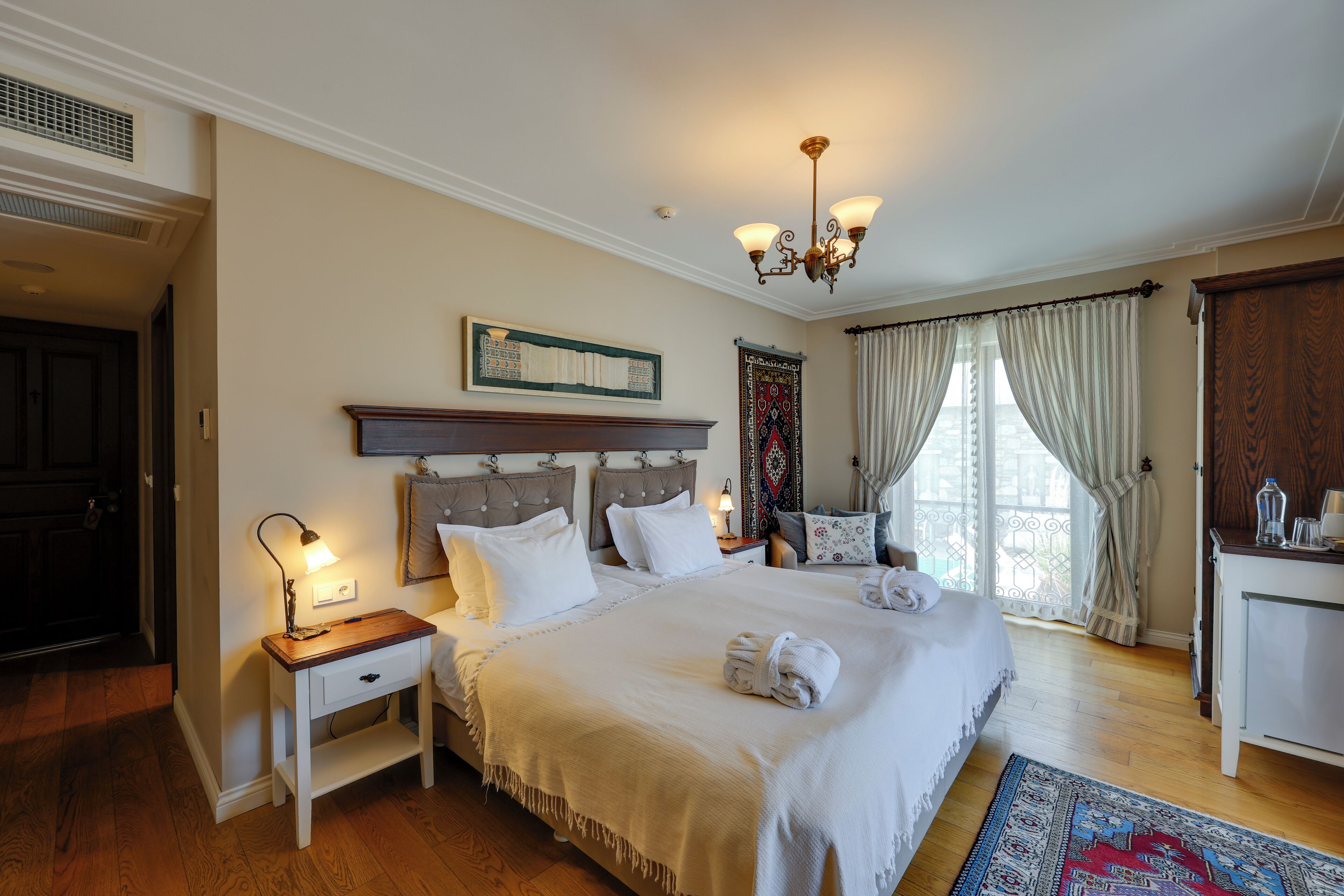Un dormitorio con una cama grande, una mesita de noche con lámpara, cabecero de madera, candelabro, ventana con cortinas y alfombra estampada.