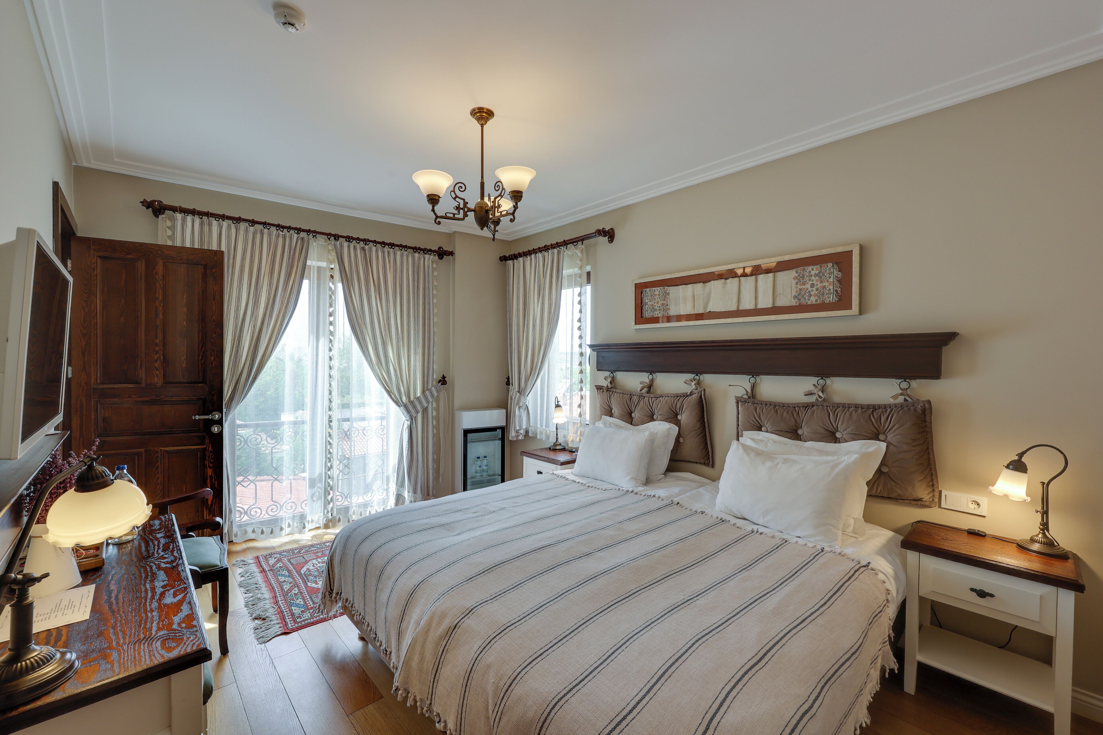A bedroom with a large bed, a desk with a lamp, a nightstand, and a chandelier.