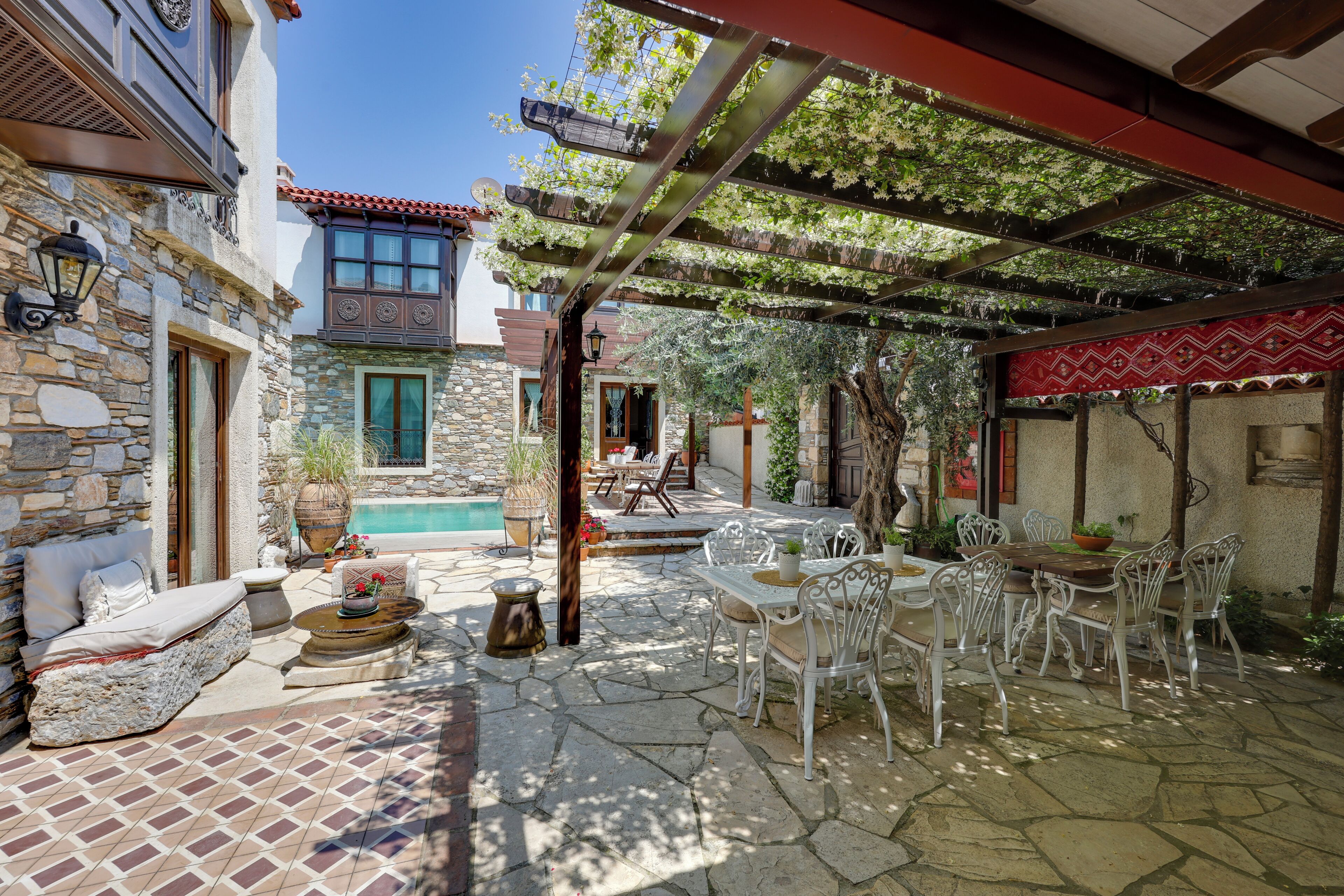 Une terrasse en pierre avec des meubles d’extérieur, une piscine et une pergola recouverte de plantes grimpantes.