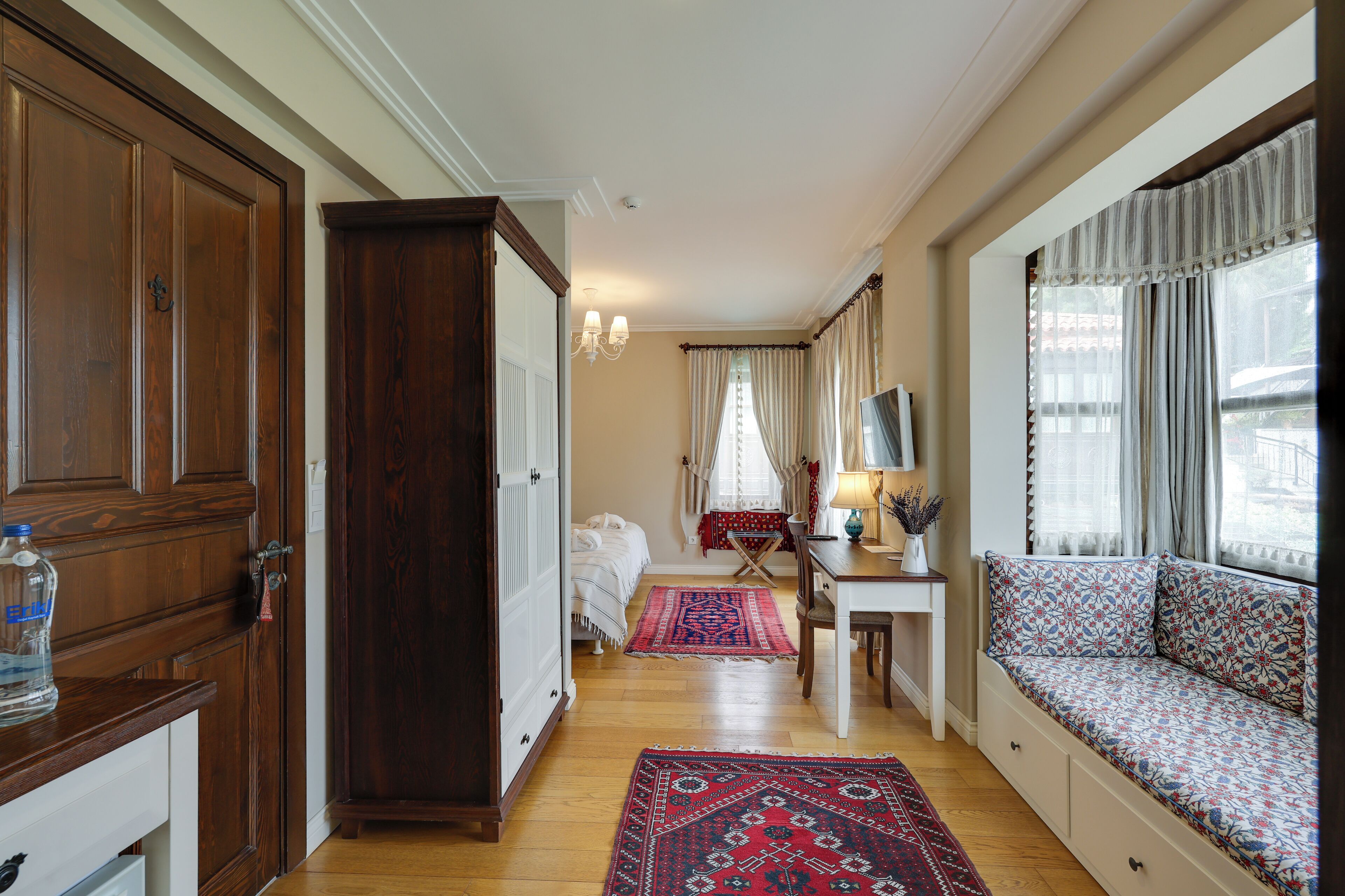Une pièce avec une armoire en bois, un lit surmonté d’un tapis rouge, un coin fenêtre agrémenté de coussins et un bureau avec une lampe.