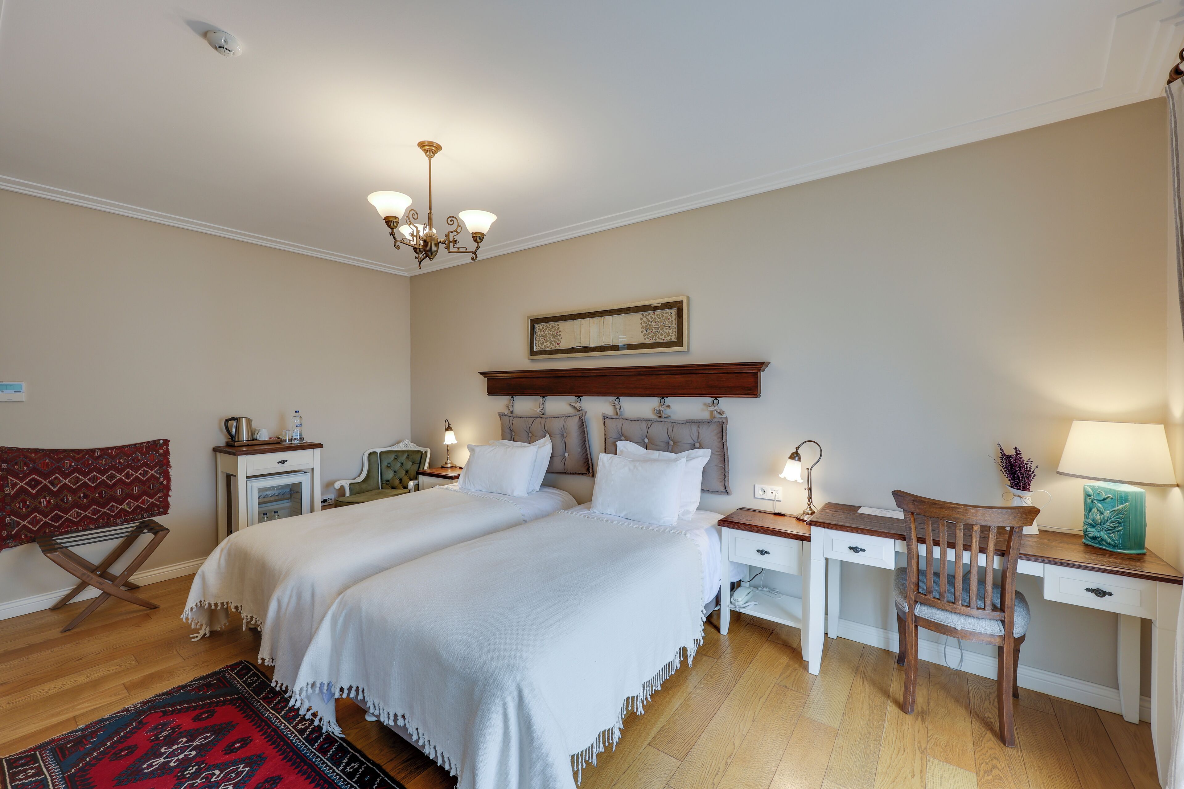 A bedroom with two beds, a desk, a chair, a chandelier, and a bedside table with lamps.