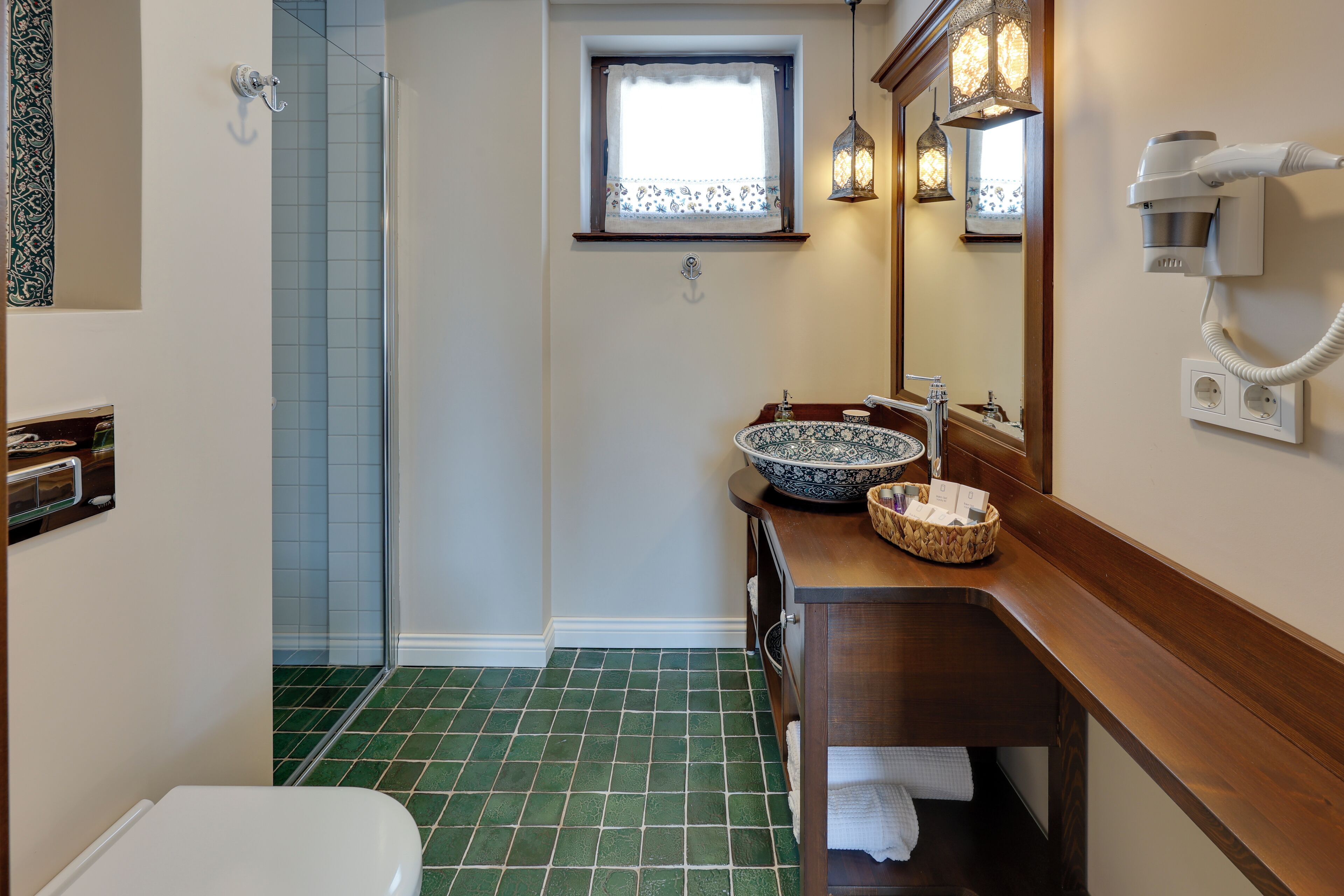 A bathroom with a green-tiled floor, a white toilet, a wooden vanity with a bowl sink, a mirror, and a hairdryer mounted on the wall.