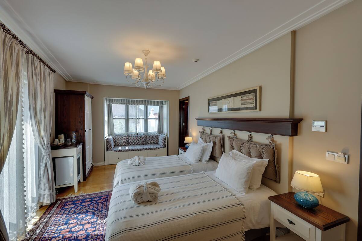 A hotel room with two beds, a desk, a window with curtains, a chandelier, and a bedside table with a lamp.