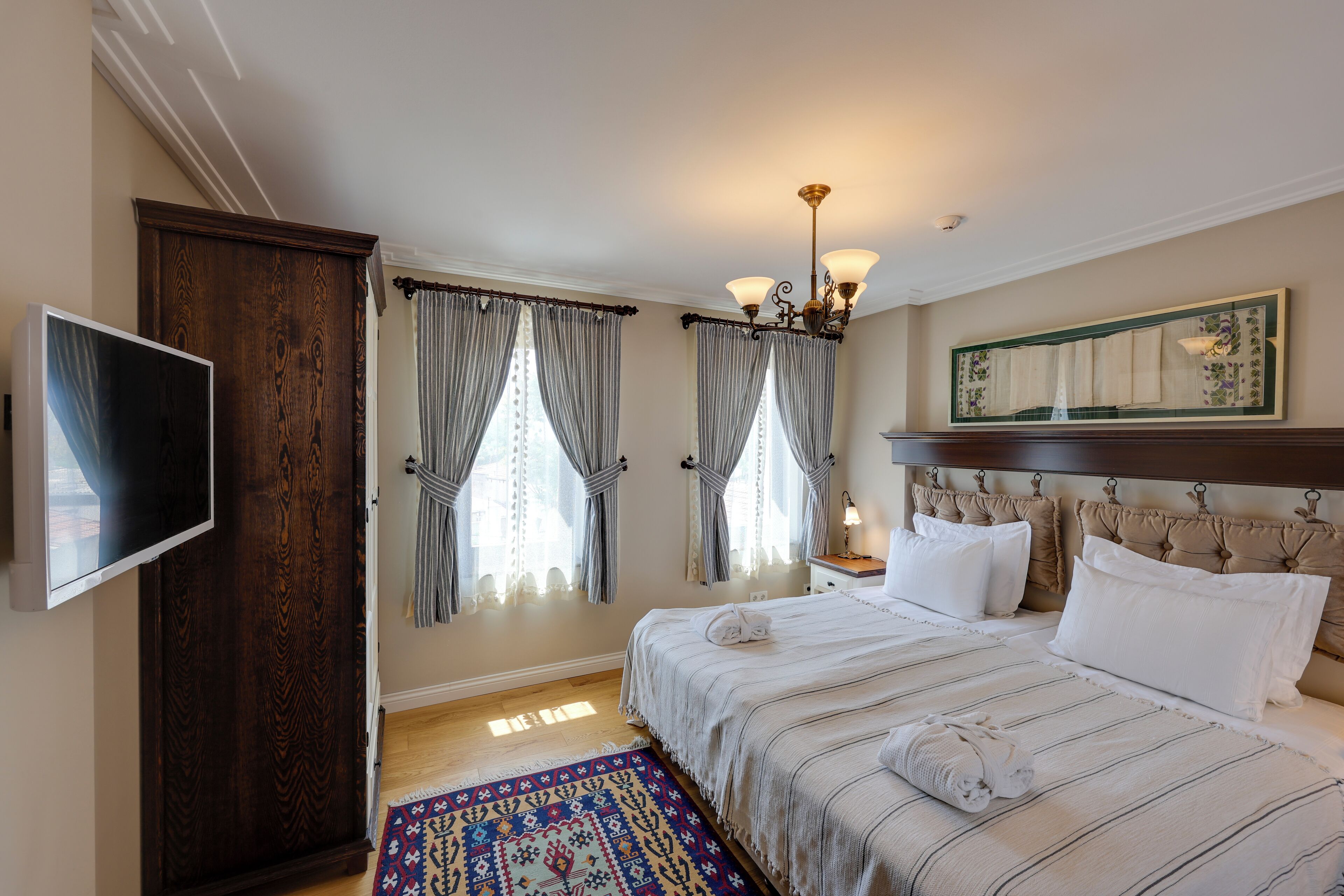 A hotel room with a bed, a television, a wooden wardrobe, a chandelier, and a patterned rug.