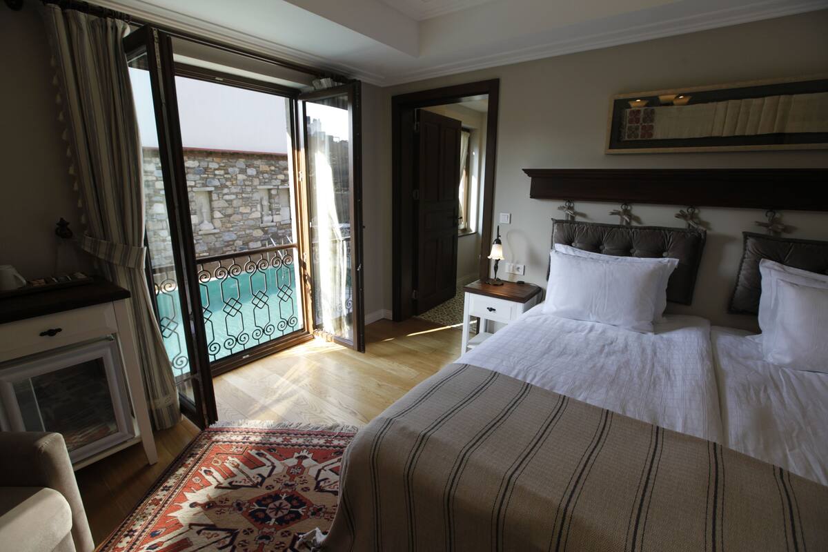 A hotel room with a bed, a desk, a chair, a small table, a nightstand, a lamp, a window with curtains, a balcony with a railing, and a patterned rug.