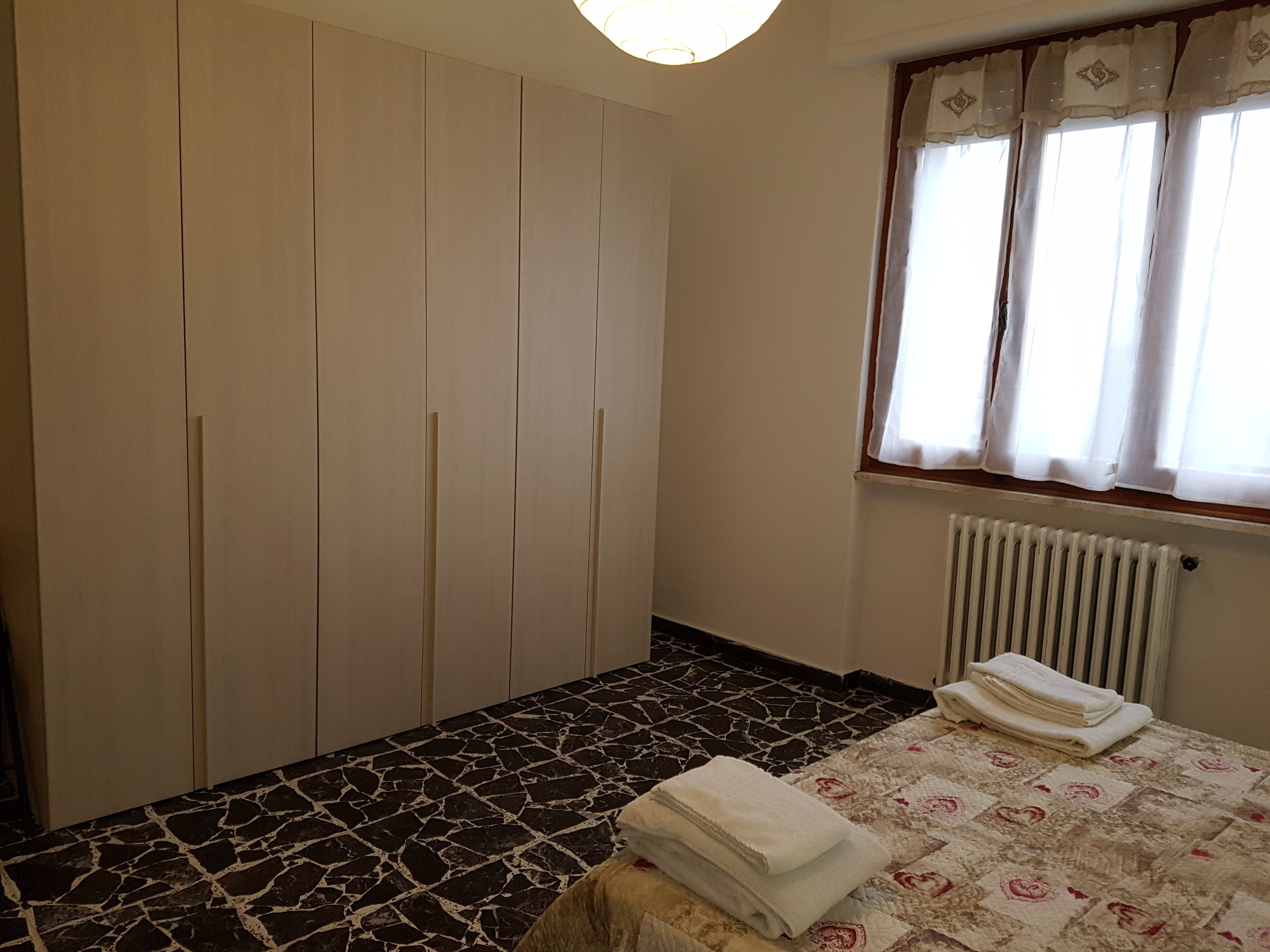 A bedroom with a bed, a radiator, a window with curtains, and a wardrobe.
