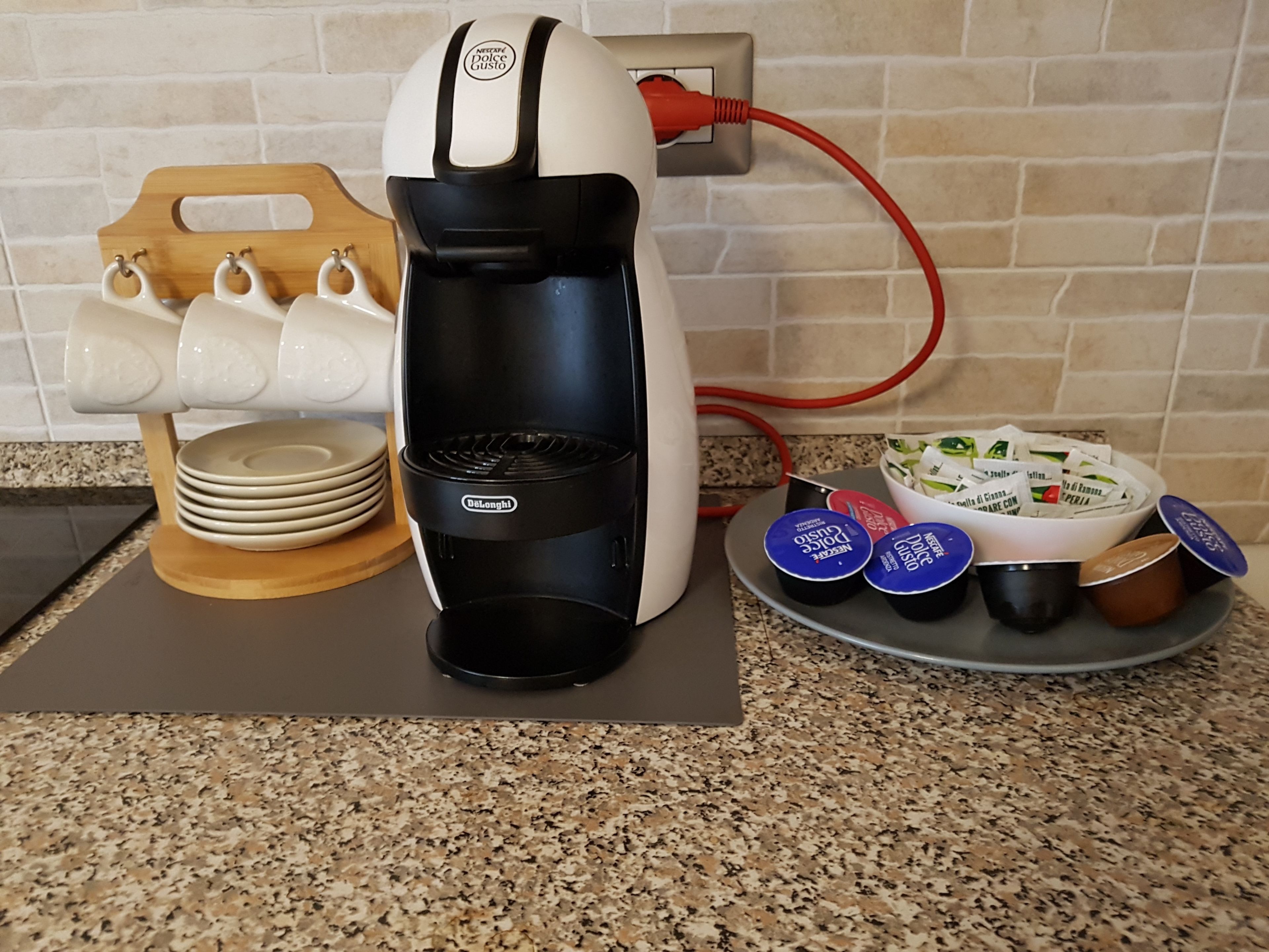 A coffee maker with a power cord plugged in, a stack of plates, and a bowl of coffee pods on a kitchen counter.
