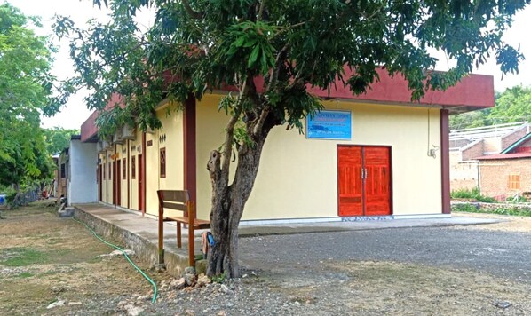 A building with a red door and a blue sign, a bench, and a tree in front. - Penginapan asidik (Bira)
