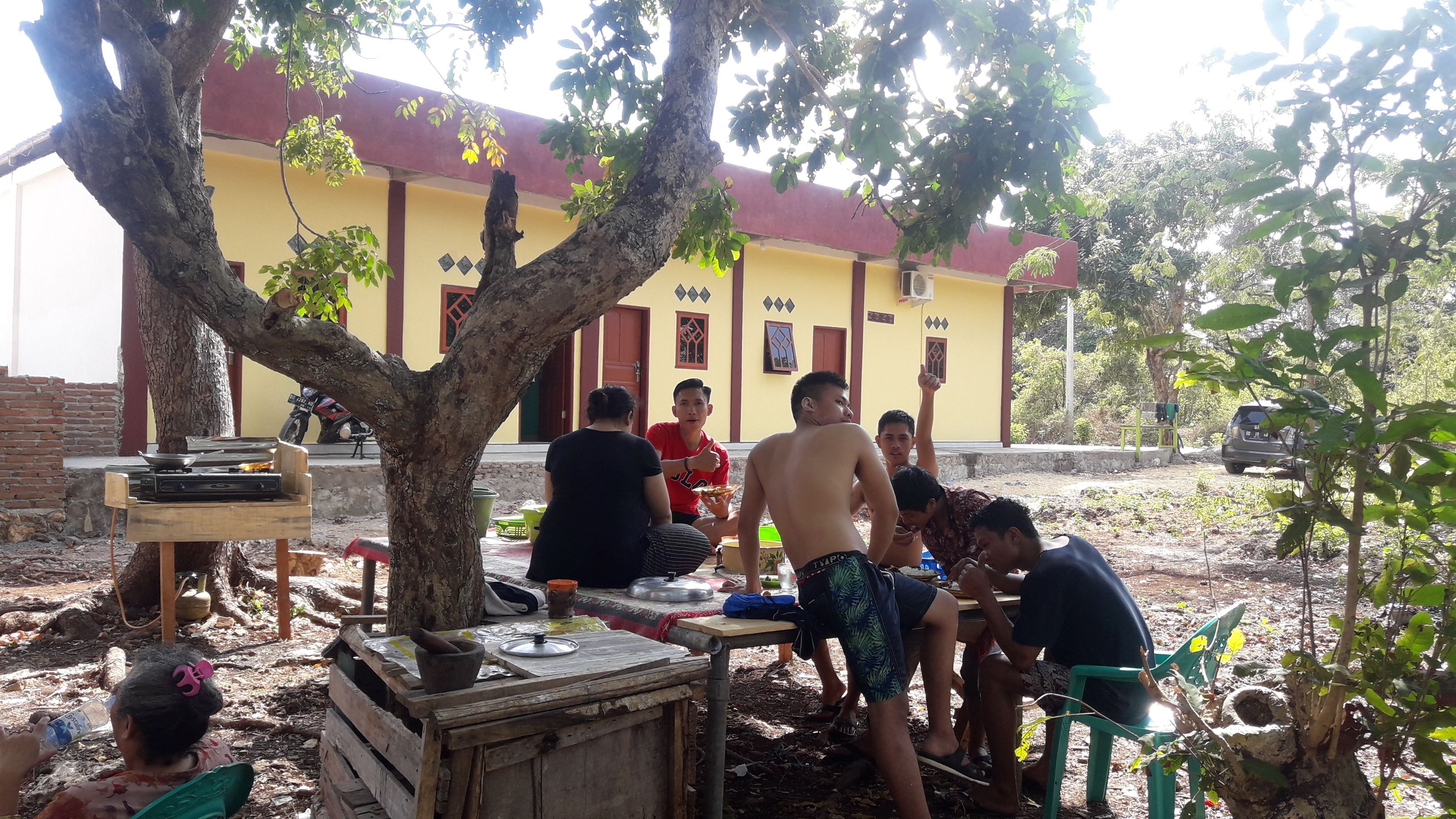 A group of people sitting outdoors under a tree, with a building in the background.