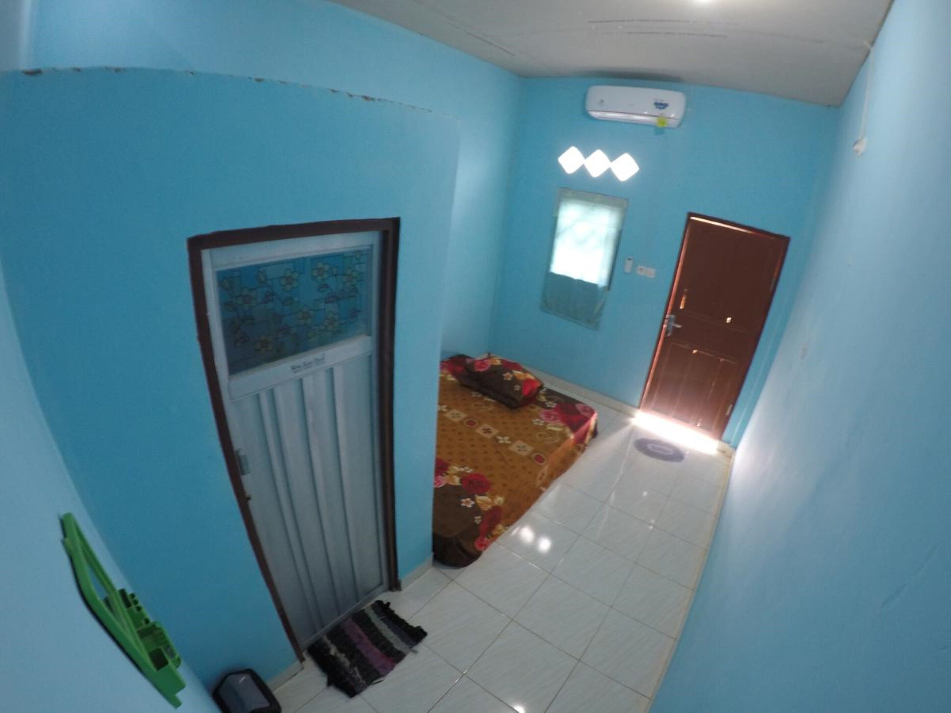 A narrow hallway with a door on the left, a window with blinds on the right, and a bed with a floral bedspread in the center.