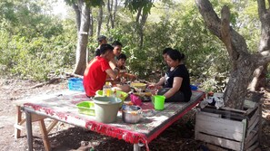 A group of people sitting at an outdoor table with various dishes and utensils, surrounded by trees. - Penginapan asidik (Bira)
