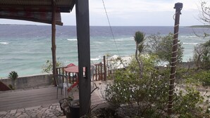 Vista al mar desde un espacio exterior cubierto con terraza de madera, un paraguas rojo y plantas en macetas.