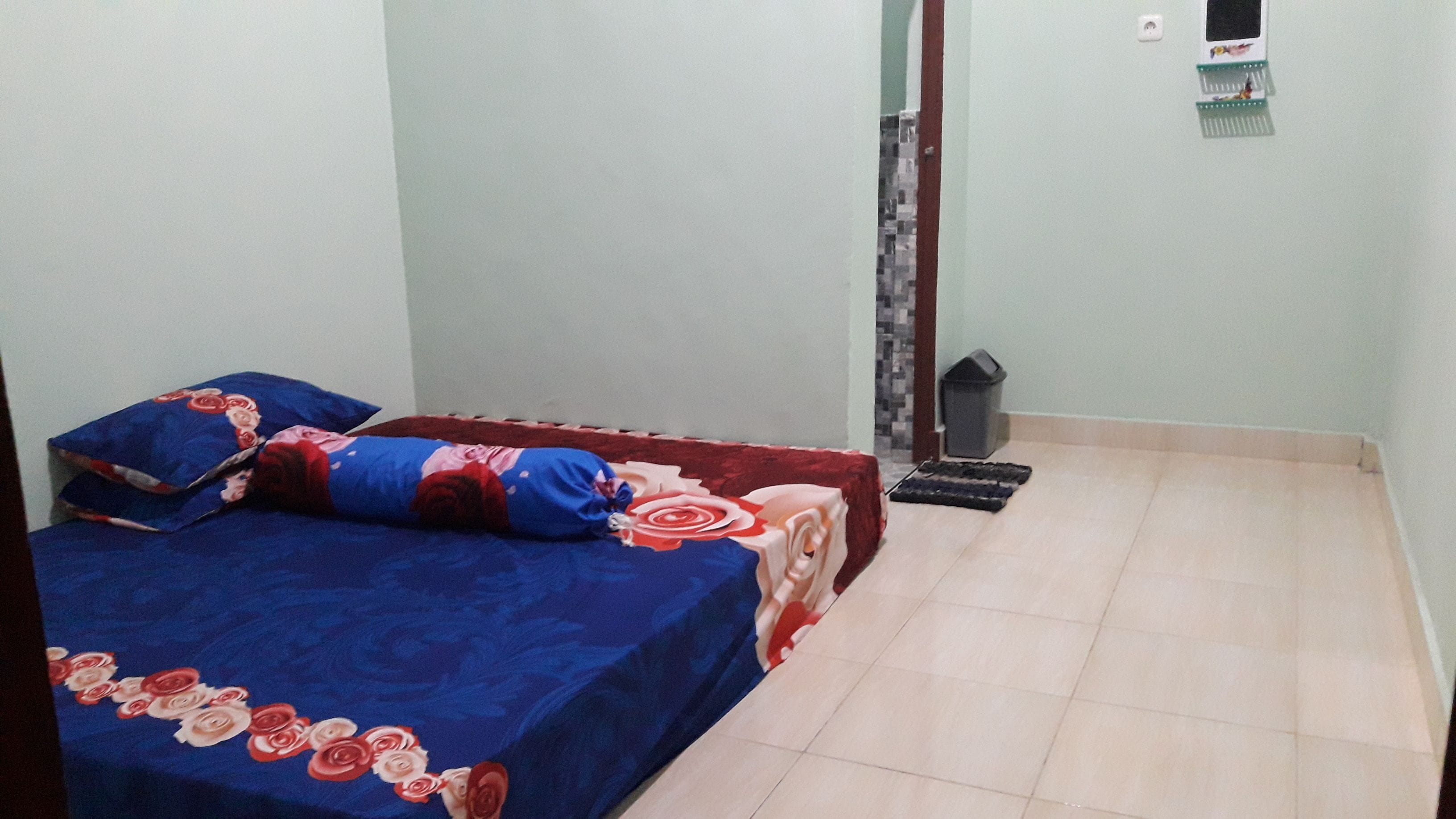 A small, simple room with a bed, a trash bin, and a wall-mounted shelf.