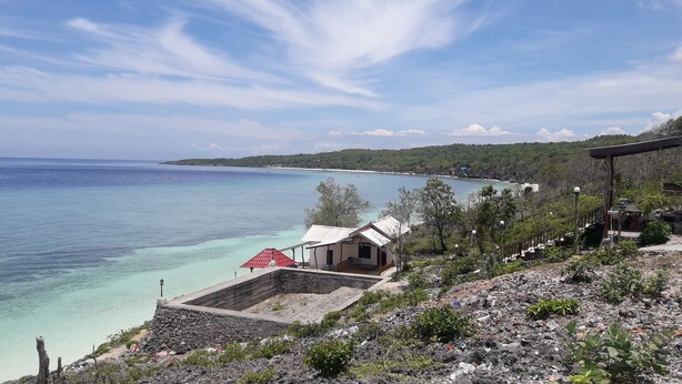 Vista costera con un edificio pequeño cerca del agua, un muro de piedra y una estructura de techo rojo.