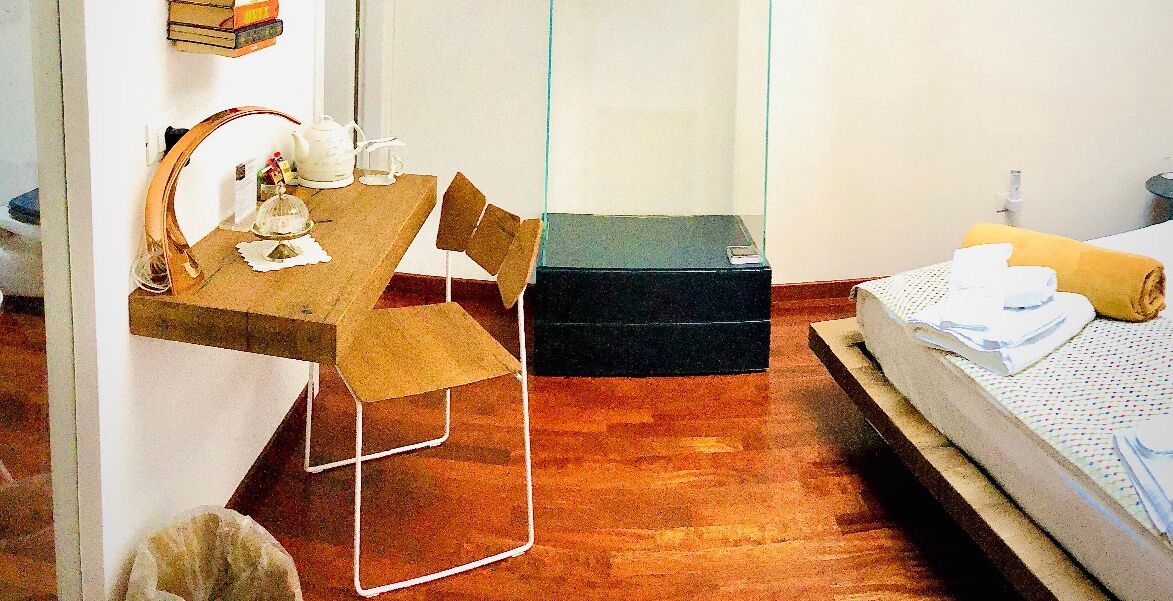 A small, compact room with a wooden floor, a simple bed with a rolled-up towel, a small table with a chair, and a few items on the table.
