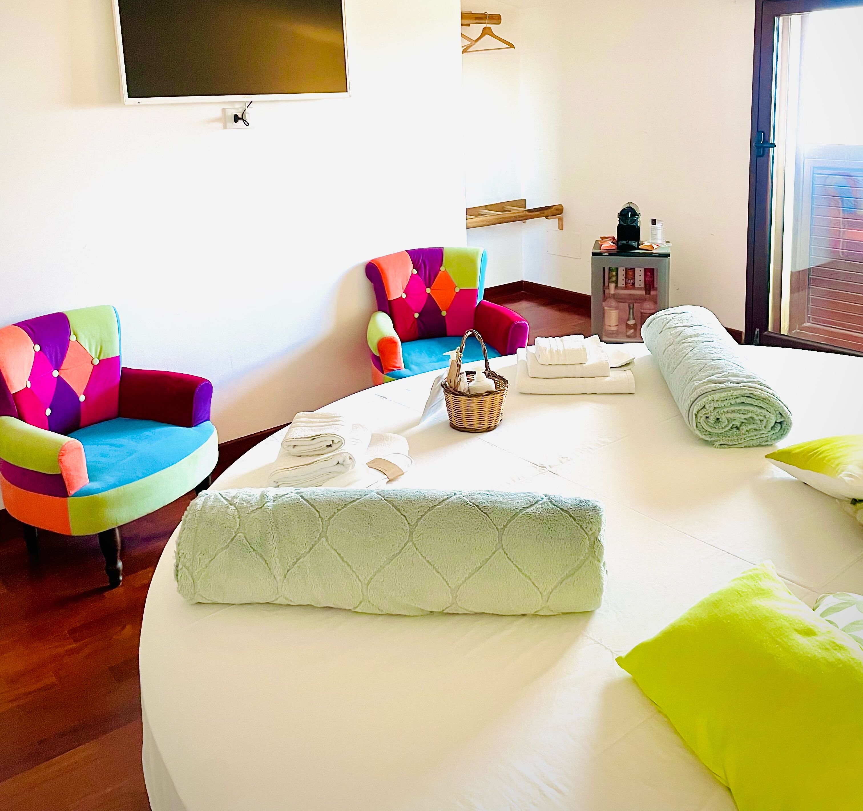 A hotel room with a bed, two colorful armchairs, a small table with a basket and towels, and a window with blinds.