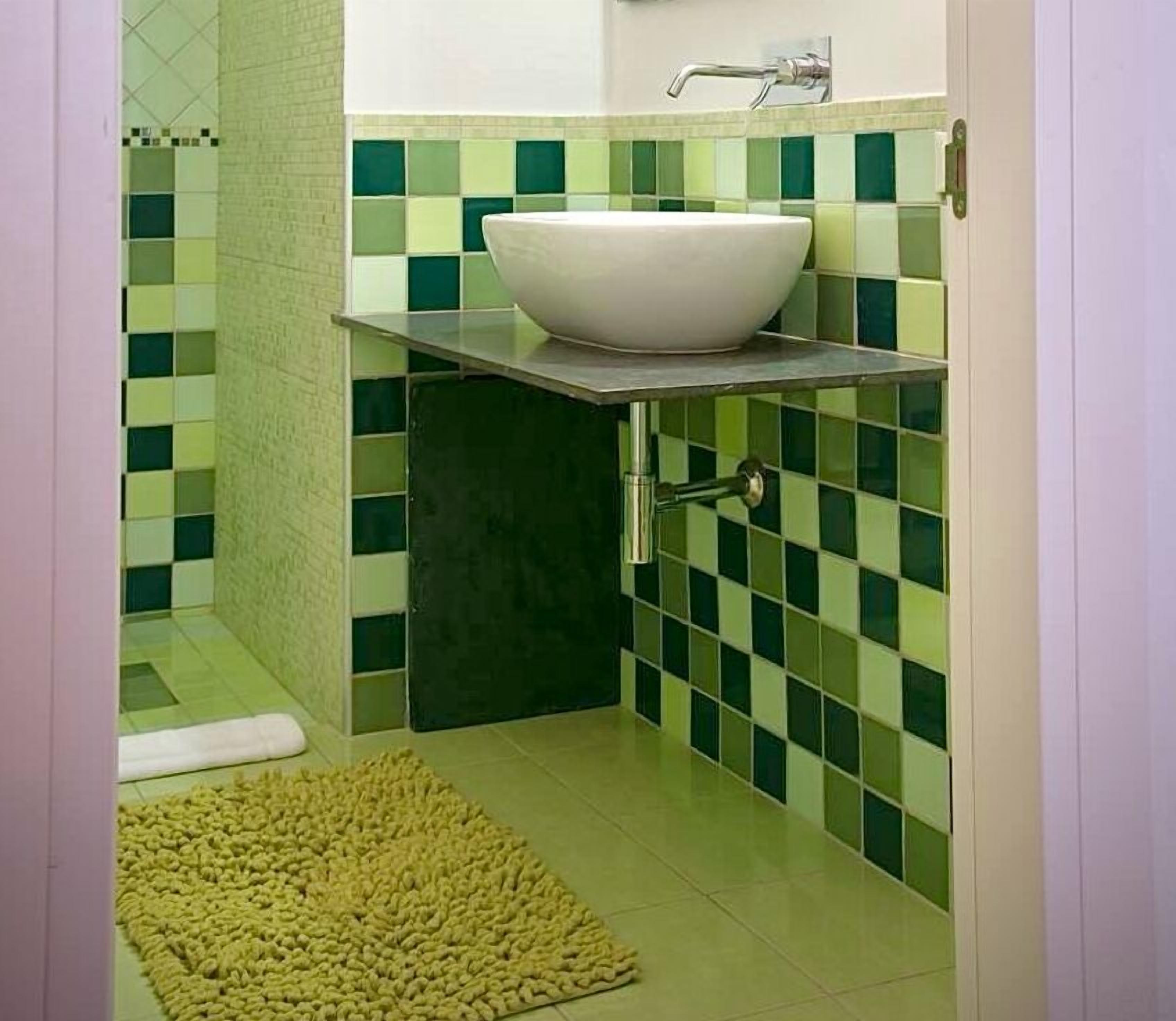 A small bathroom with a white sink, green and white tiles, and a yellow rug on the floor.