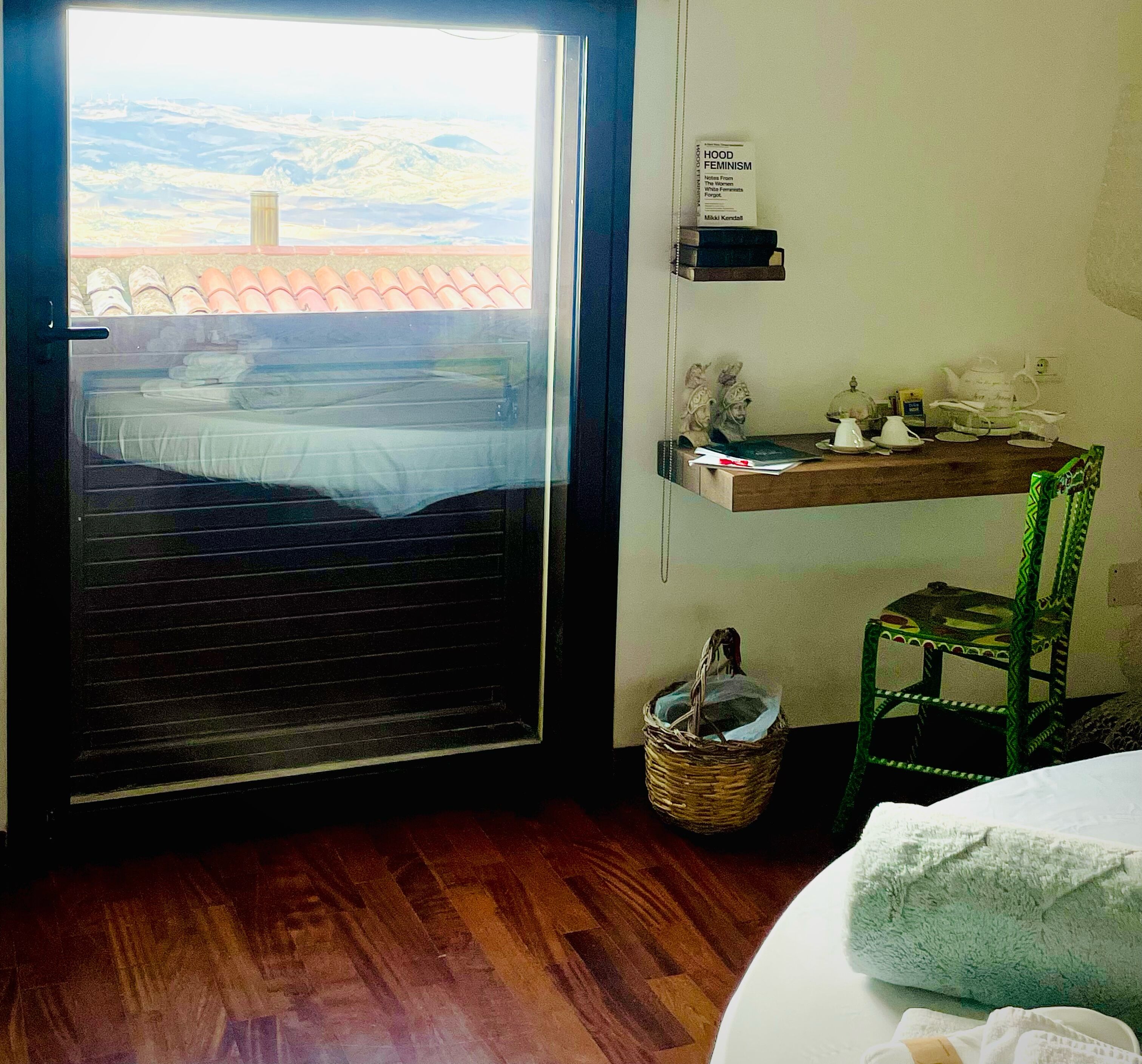 A room with a wooden floor, a bed with a white cover, a green chair, a small table with a book and a teapot, and a window with a view of hills and a red-roofed building.