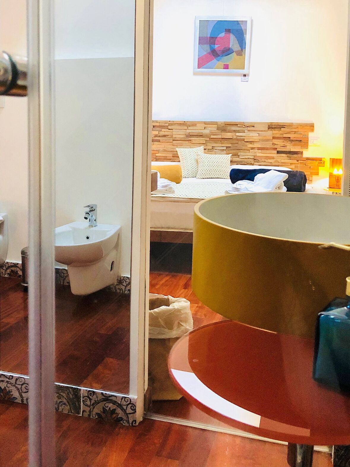 A hotel room with a wooden headboard, a bed with white and patterned bedding, a bedside table with a lamp, and a bathroom with a sink and toilet.