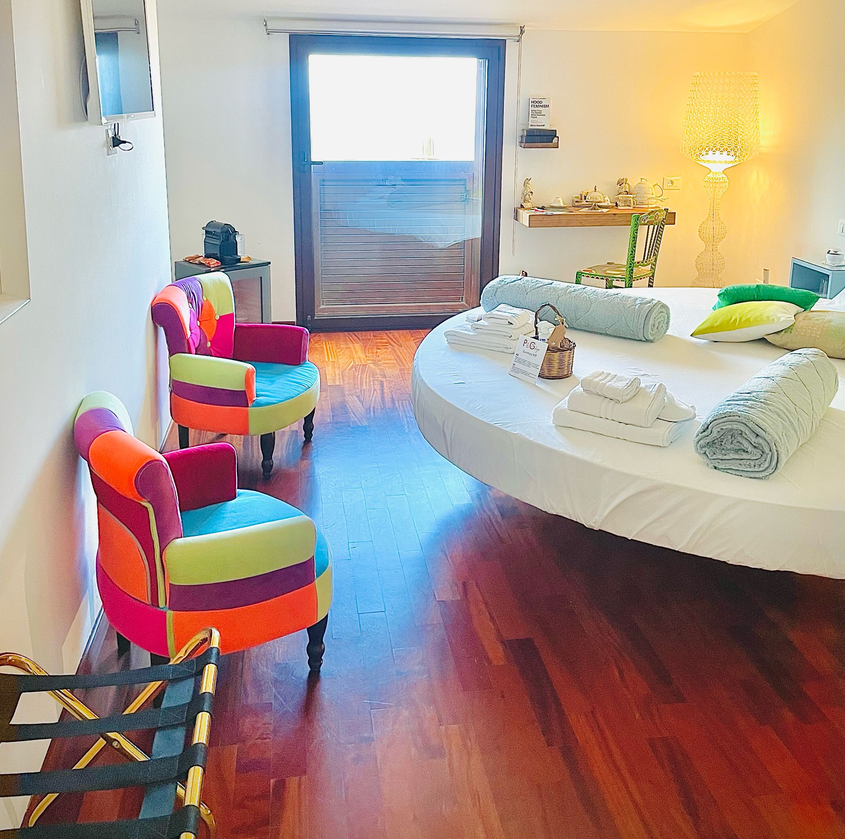 A hotel room with a round bed, colorful chairs, a small table with toiletries, and a view through a large window.