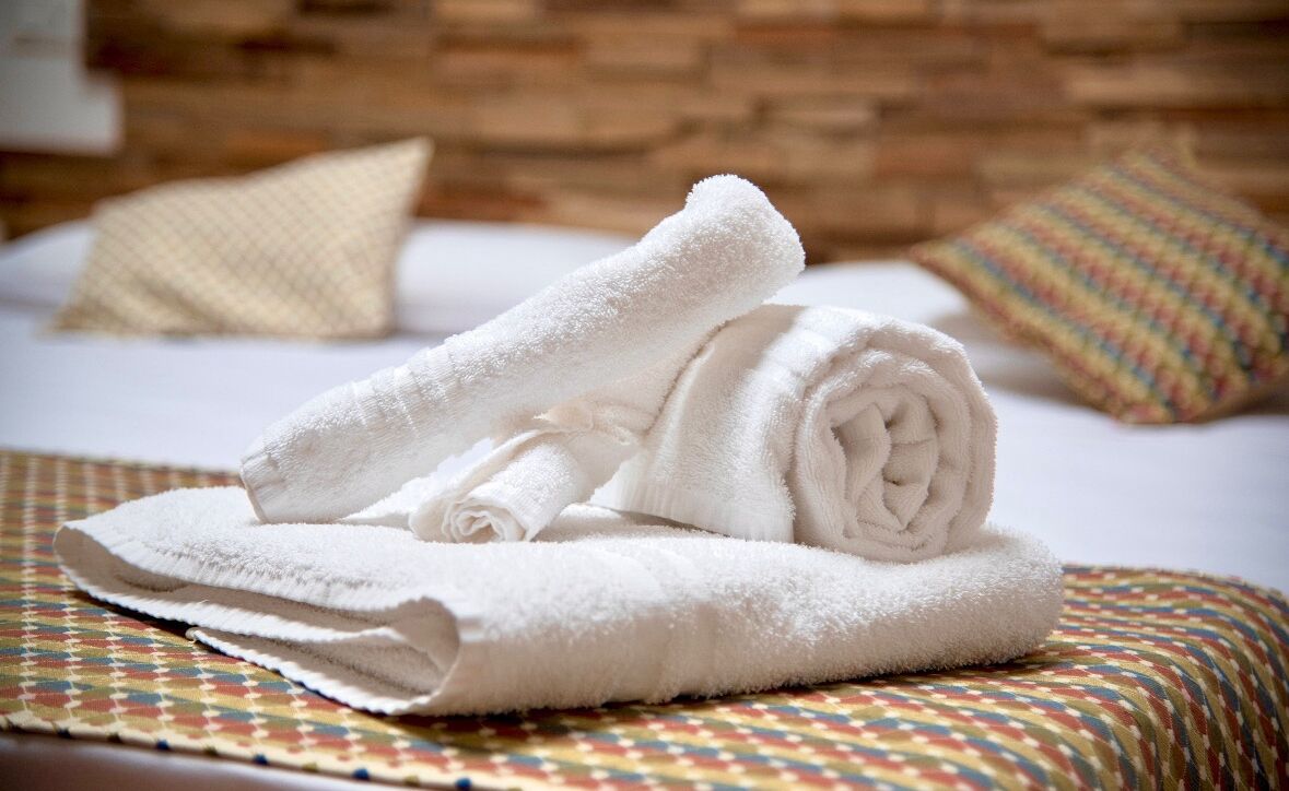 A neatly folded white towel on a bed with a patterned pillow and a plain pillow in a hotel room.