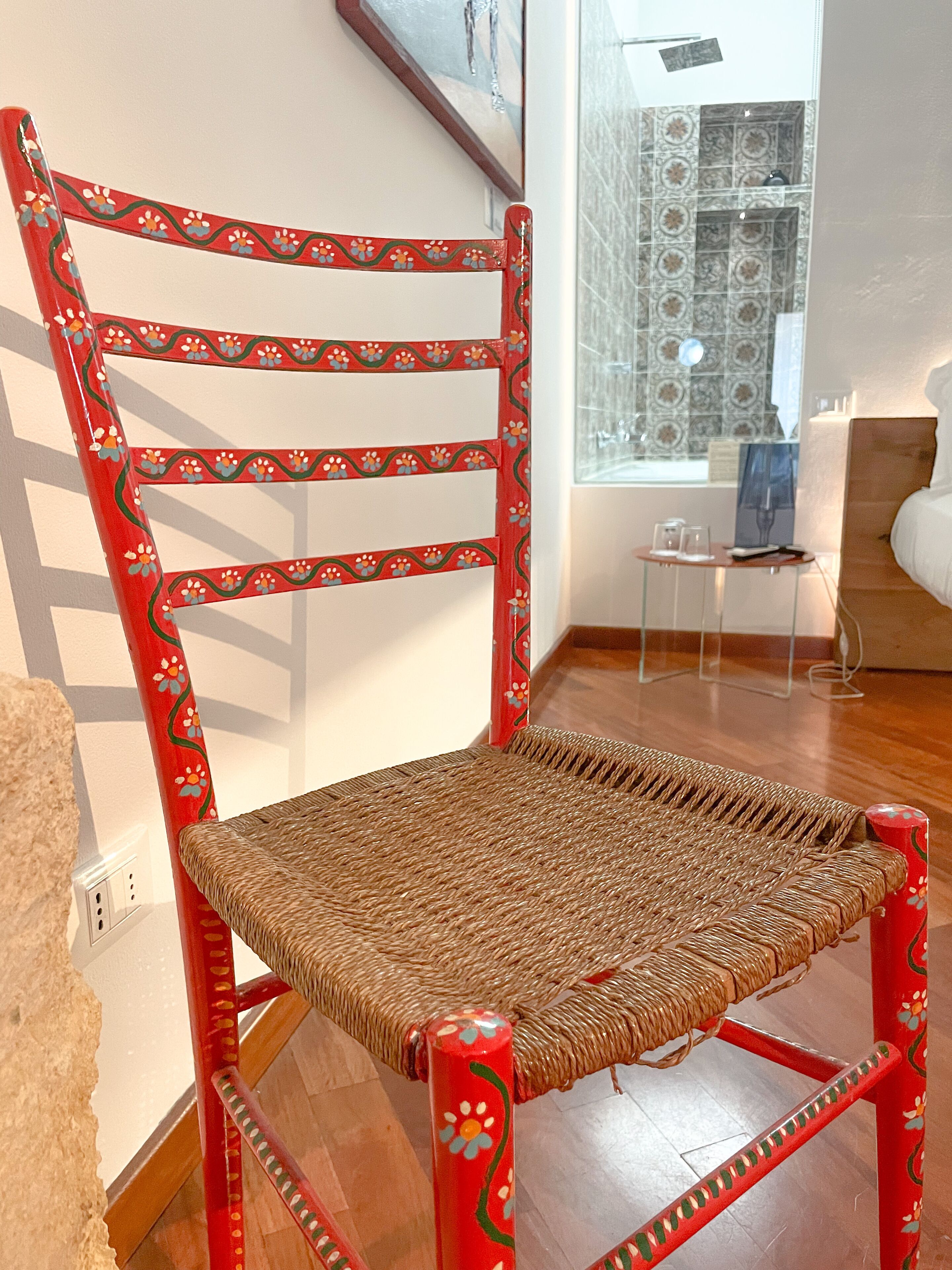 A red chair with a woven seat and backrest, positioned in a room with a wooden floor and a window with patterned curtains.