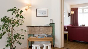 Una habitación con una mesa de madera, dos taburetes, una planta en maceta, un cuadro en la pared y un sofá rojo al fondo.