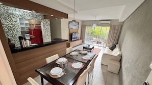 A modern dining area with a long table set for four, a kitchen with a mosaic backsplash, and a living area with a sofa and a TV. - Beach Living, Frente mar, 400m do B Park (Aquiraz)
