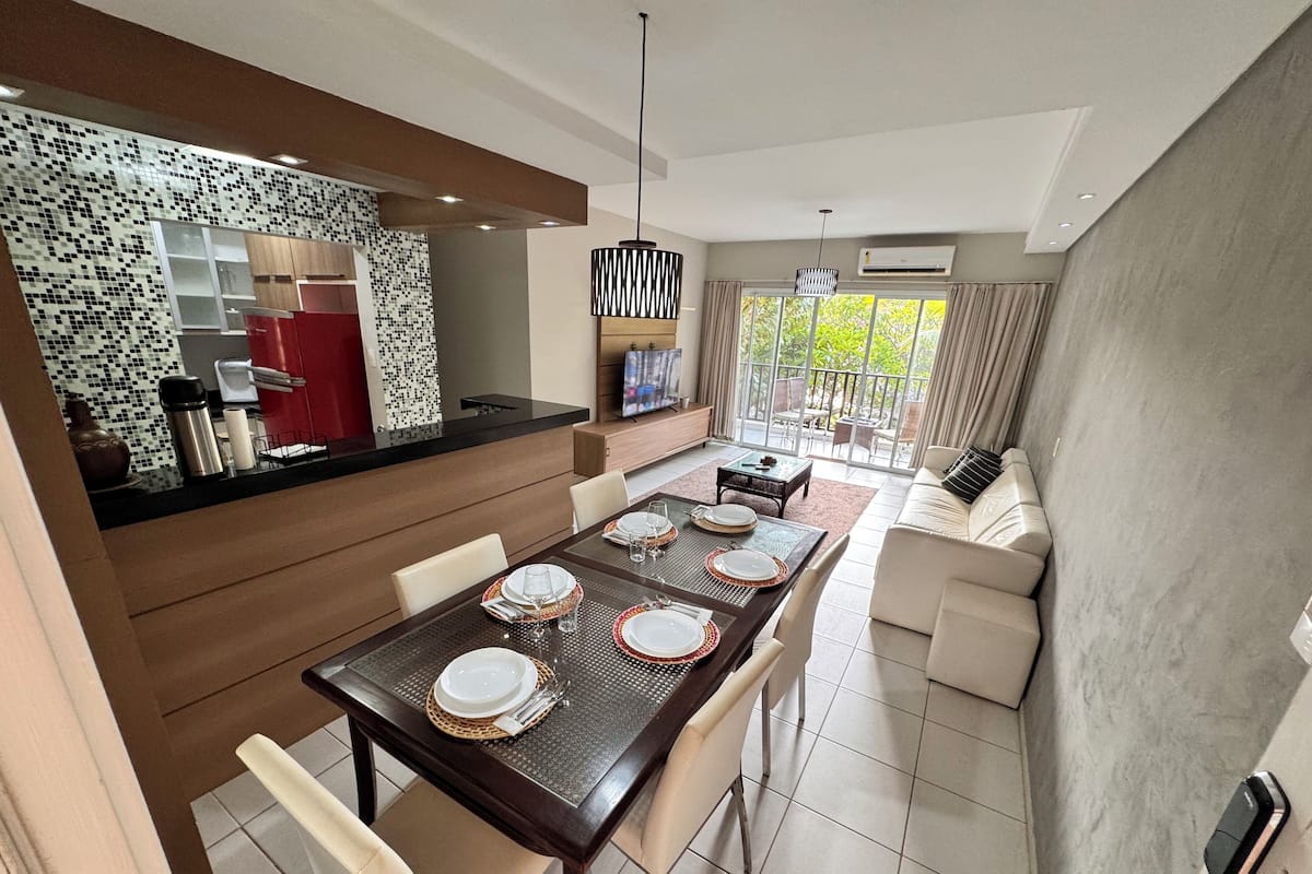 A modern dining area with a long table set for four, a kitchen with a mosaic backsplash, and a living area with a sofa and a TV.