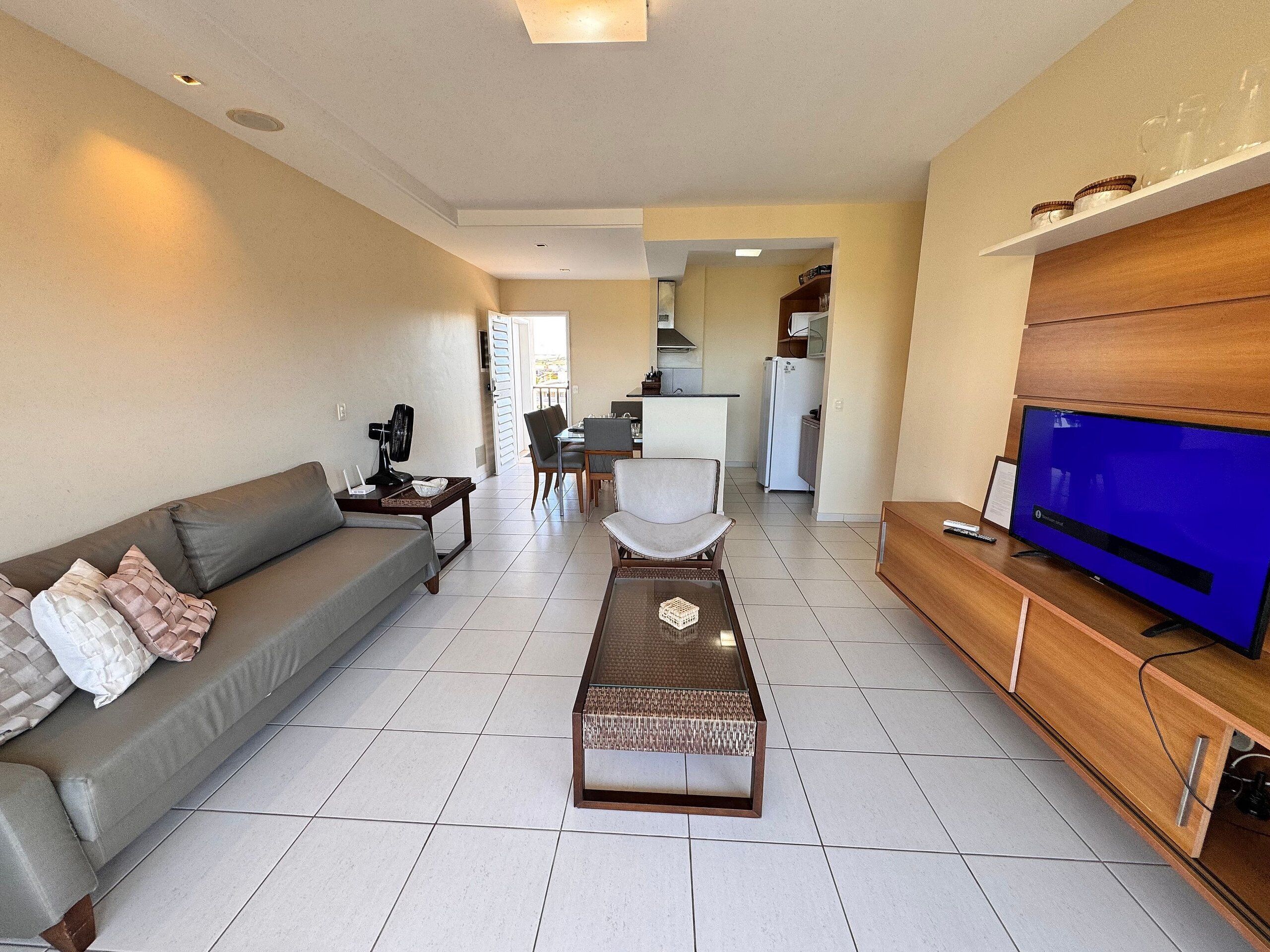 Una sala moderna con un sofá, una mesa de centro, un televisor y una cocina al fondo.
