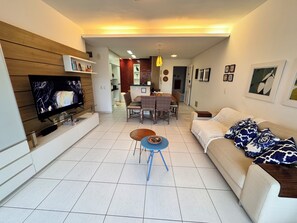 A modern living room with a flat-screen TV, a sofa, a coffee table, and a dining area with chairs and a table. - Beach living, Pé na areia, Vizinho ao B Park - Oficial (Aquiraz)
