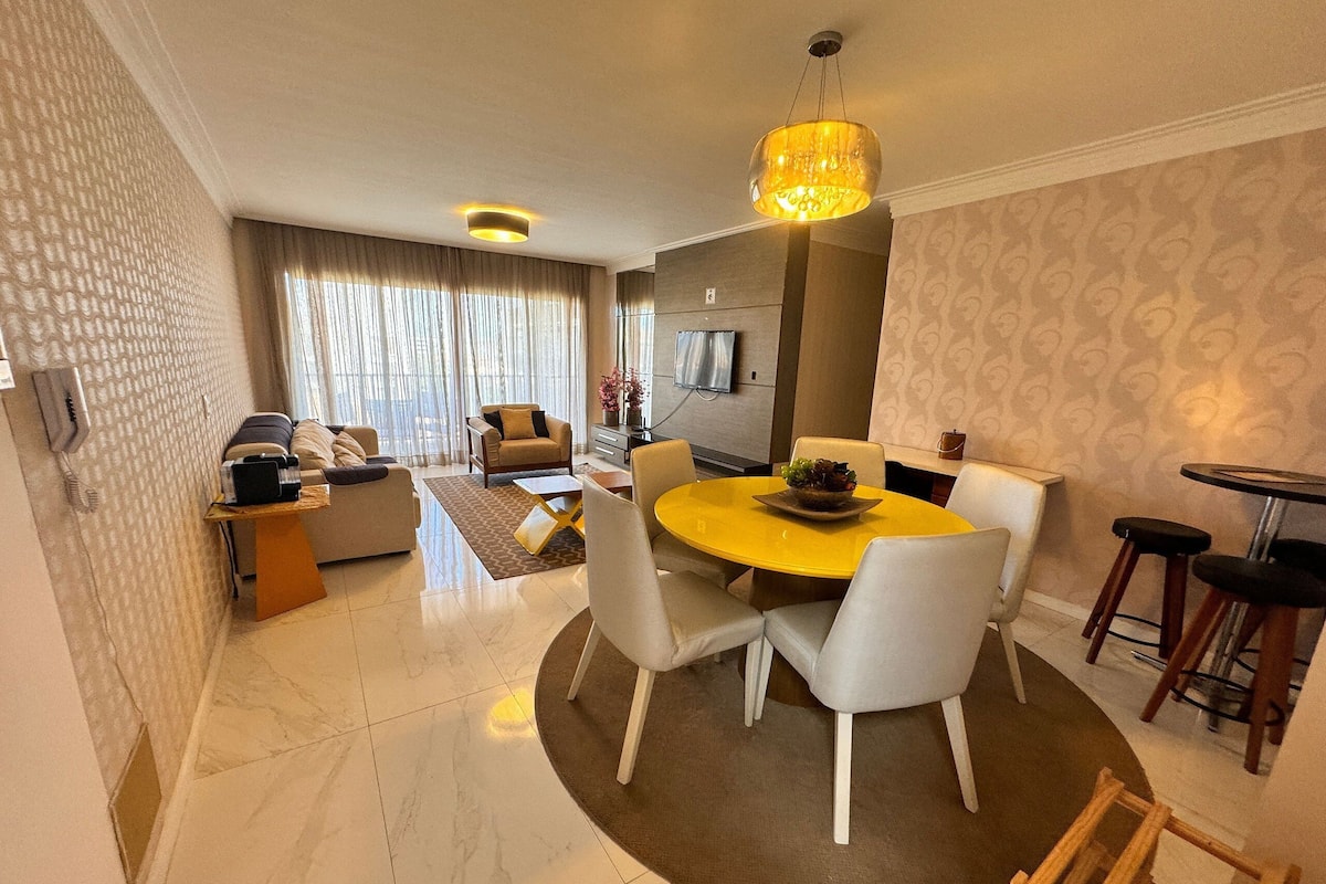 A modern living room with a dining area, featuring a round yellow table, white chairs, a sofa, and a television.