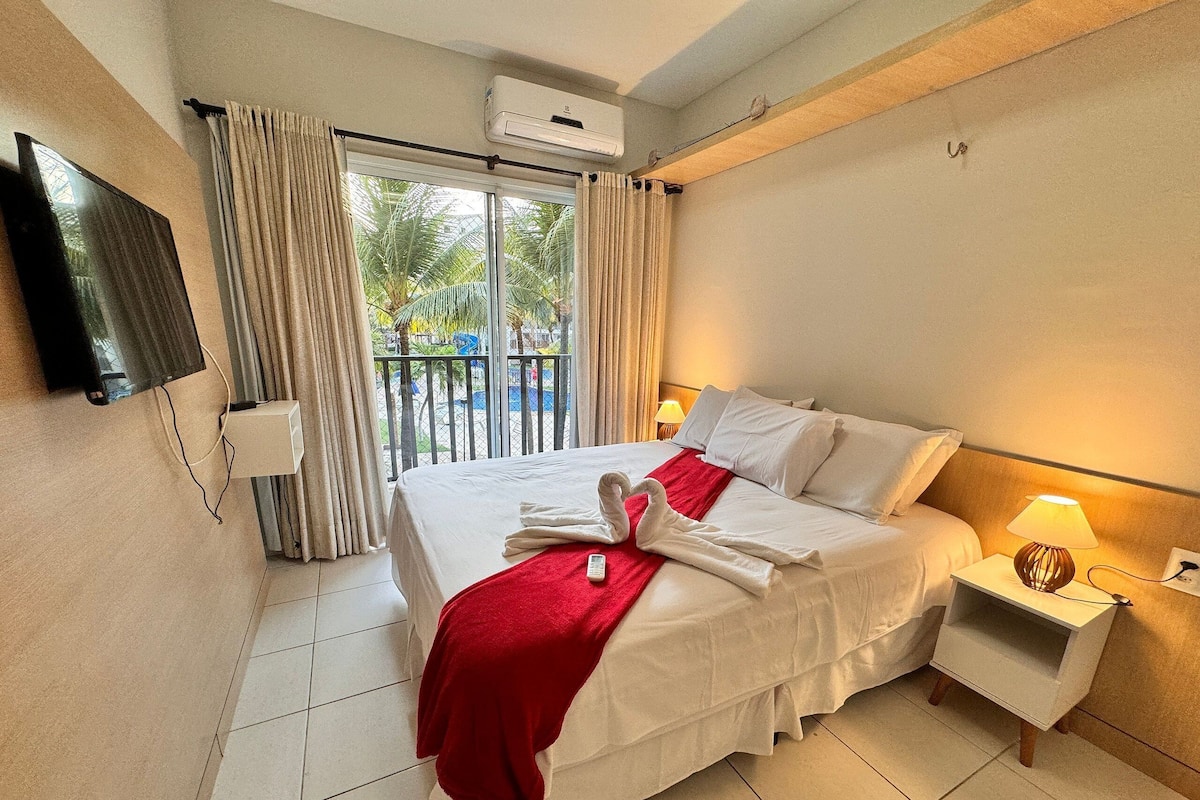 A hotel room with a bed, a TV mounted on the wall, a bedside table with a lamp, and a view of the outdoors through the window.