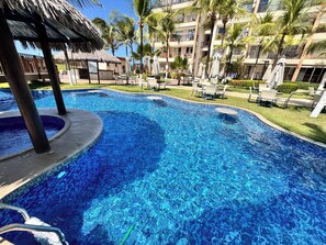 A swimming pool with a thatched-roof structure, surrounded by palm trees and lounge chairs. - Beach living, Pé na areia, Vizinho ao B Park - Oficial (Aquiraz)