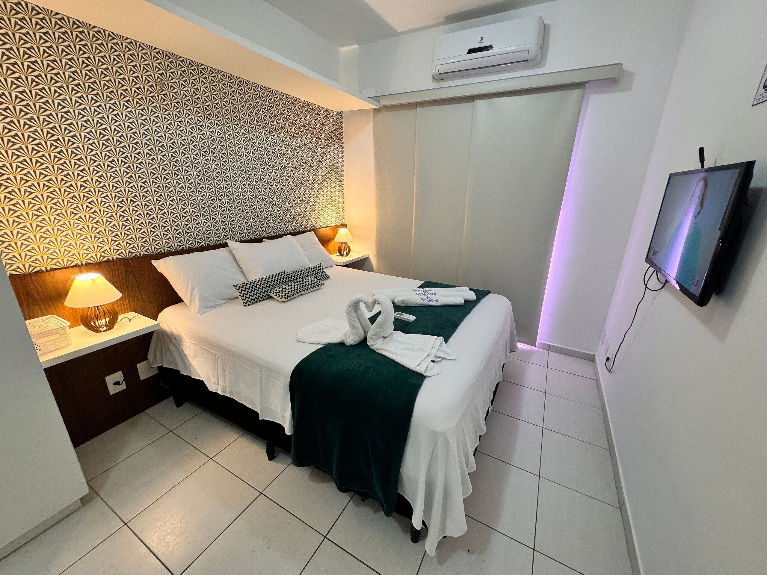 A hotel room with a patterned wallpaper, a bed with white linens, a green throw blanket, and a flat-screen TV mounted on the wall.