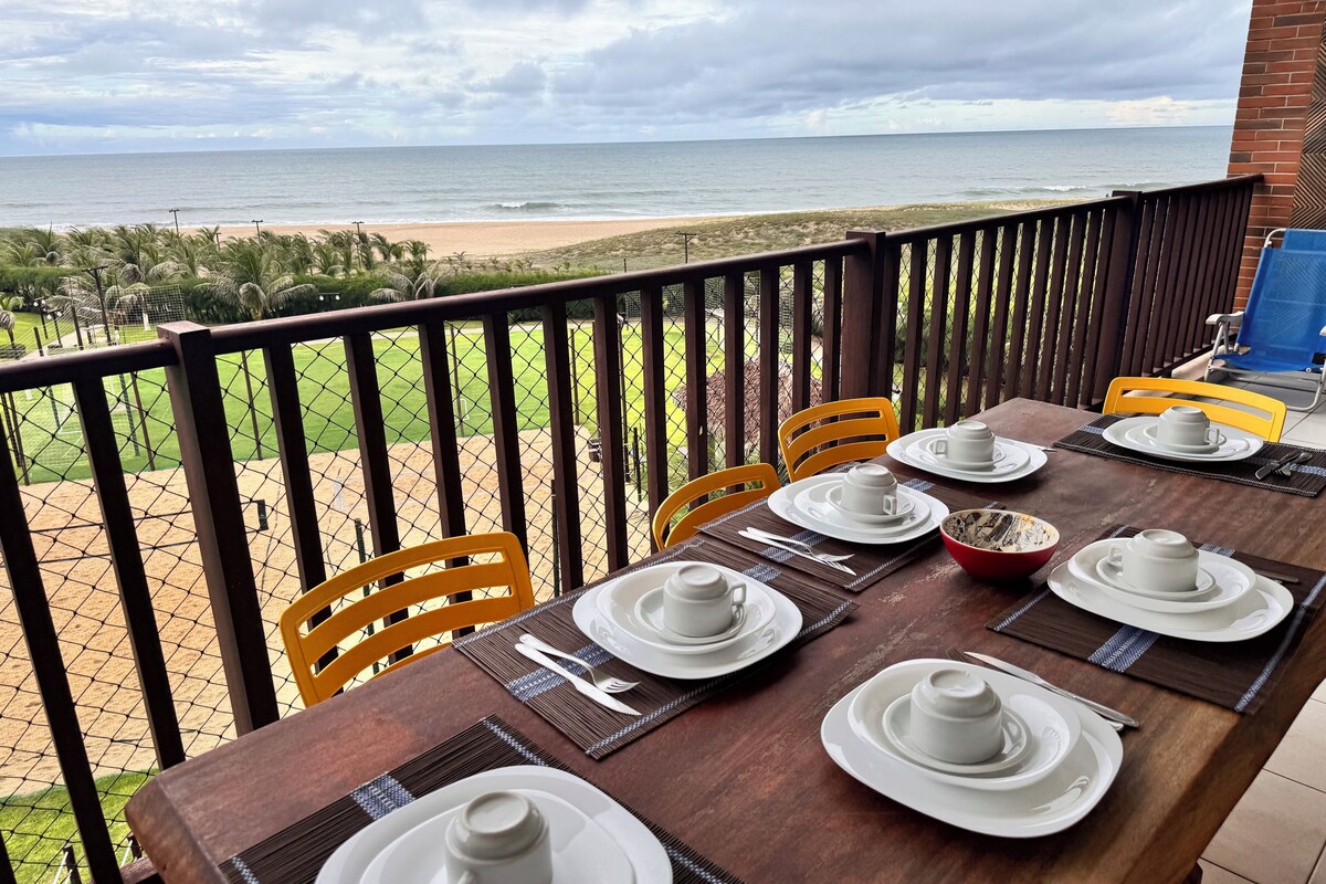 A dining table set for four with white plates and cups, overlooking a beach and ocean.
