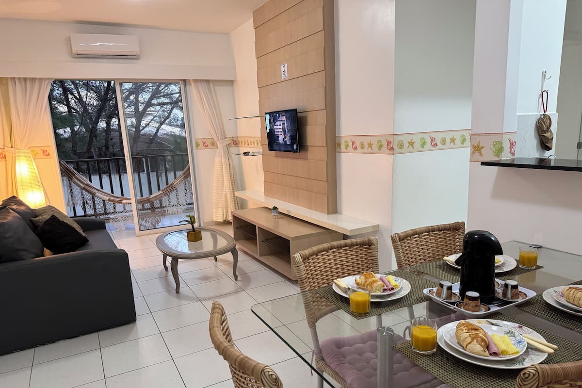 A modern living room with a sofa, a glass coffee table, wicker chairs, a TV mounted on the wall, and a balcony with a view of trees.